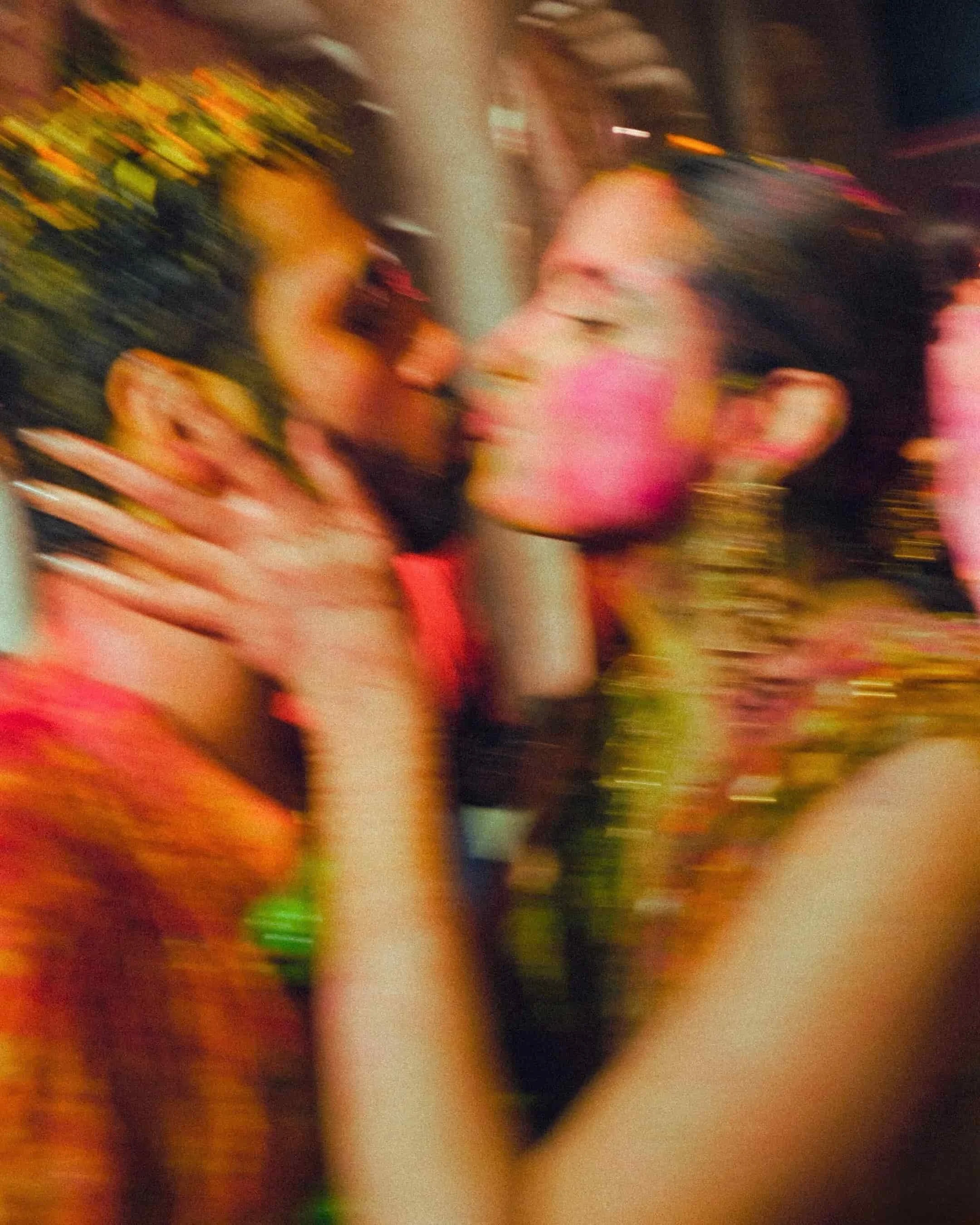 Two women are close together, smiling and possibly about to kiss. The women are wearing traditional or brightly colored clothing with jewelry and flowers, indicating a festive or cultural celebration.