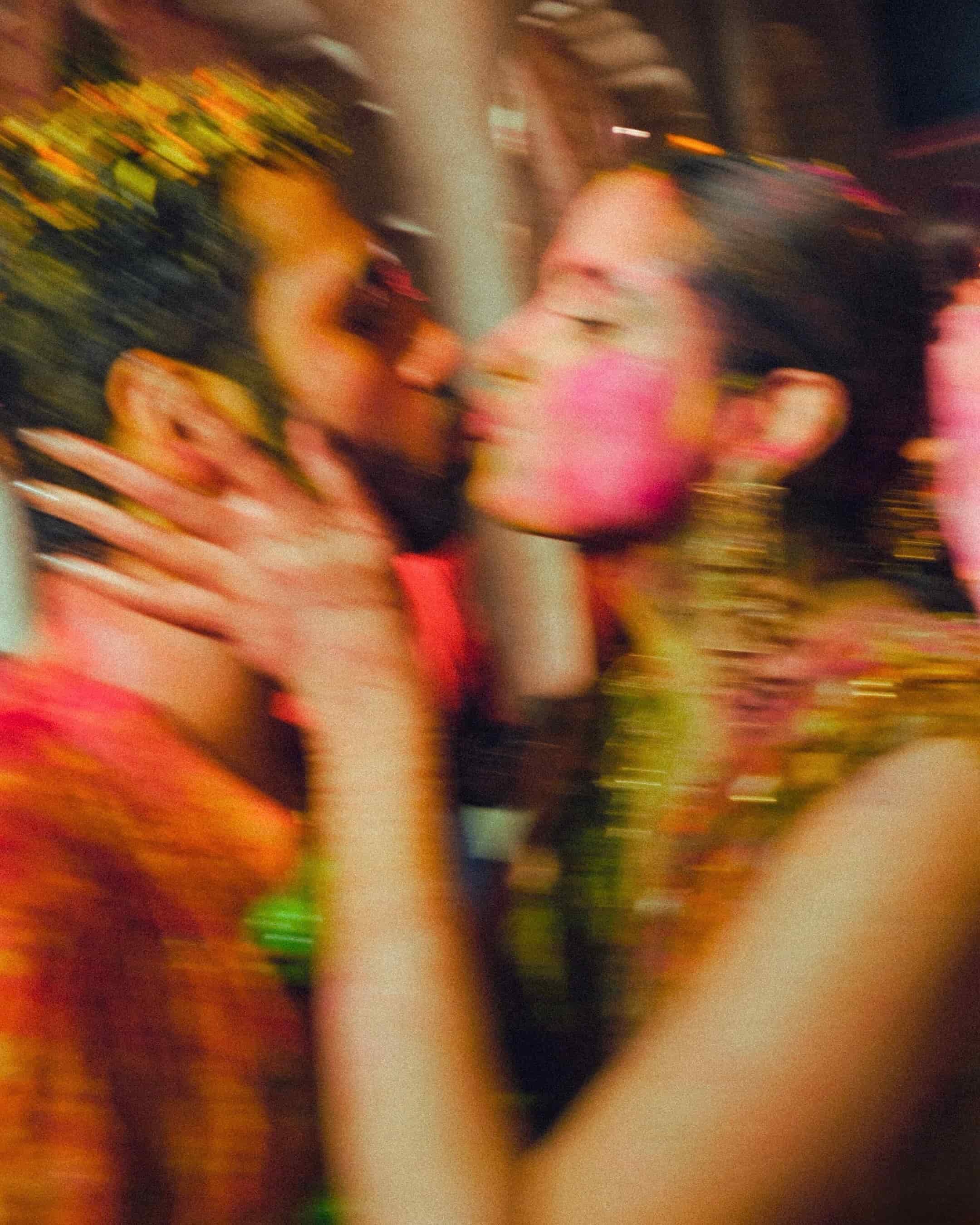 Two women dressed in traditional Indian wedding attire, close to each other, smiling and about to kiss, with vibrant jewelry, makeup, and colorful background.