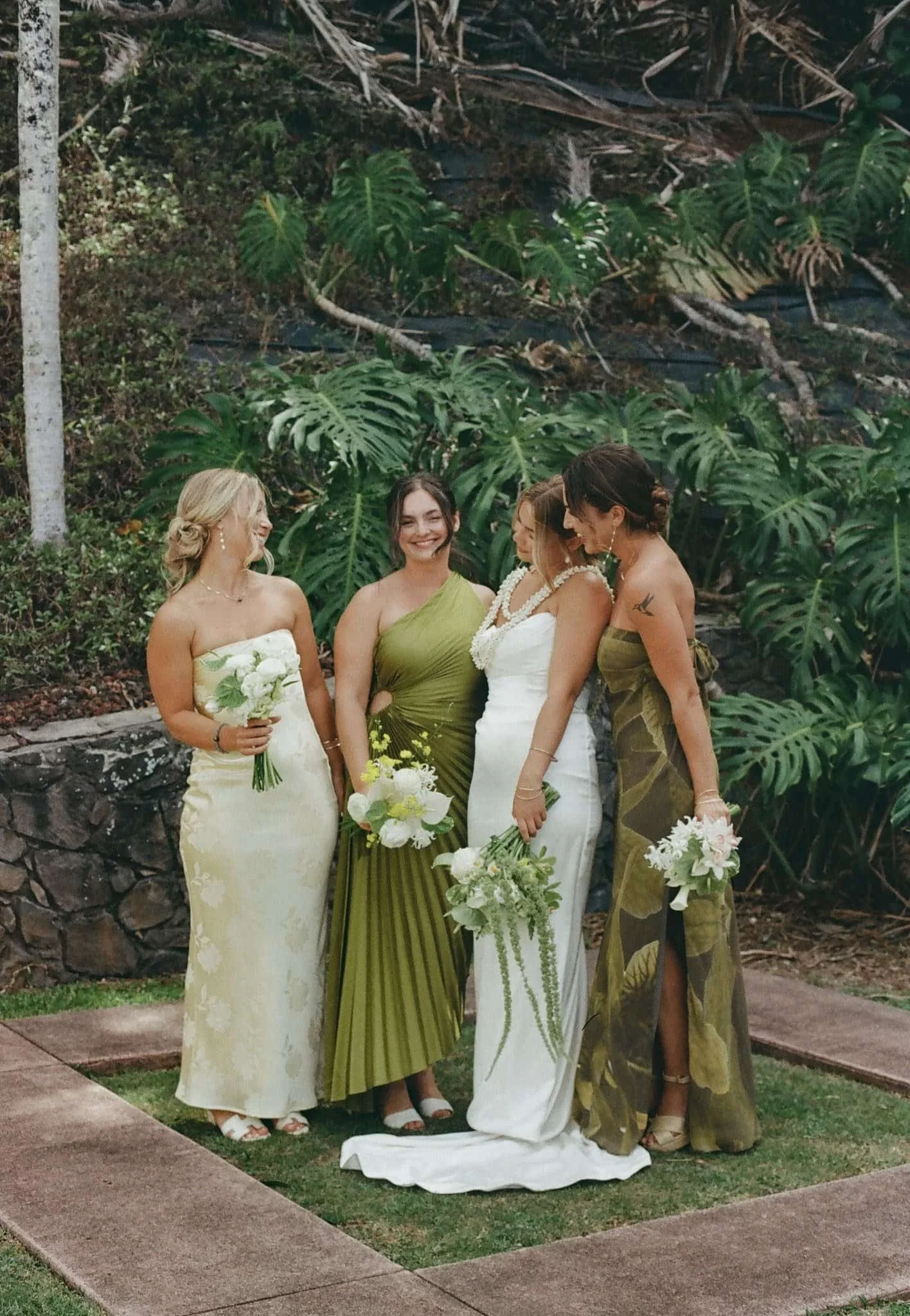 Four women dressed in elegant dresses, standing in a garden with large green tropical plants, celebrating a wedding.