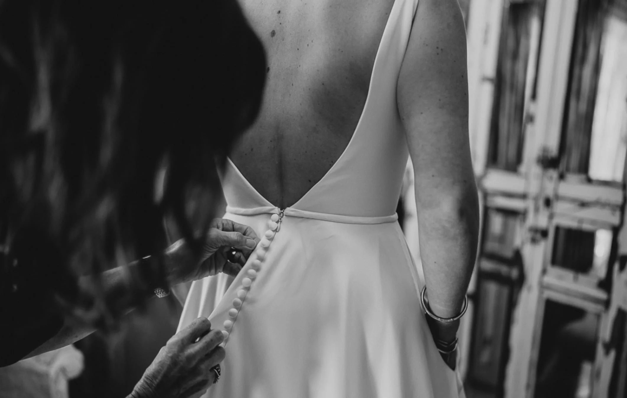 A person is adjusting buttons on the back of a woman wearing a white dress, likely in a fitting or dressing room.