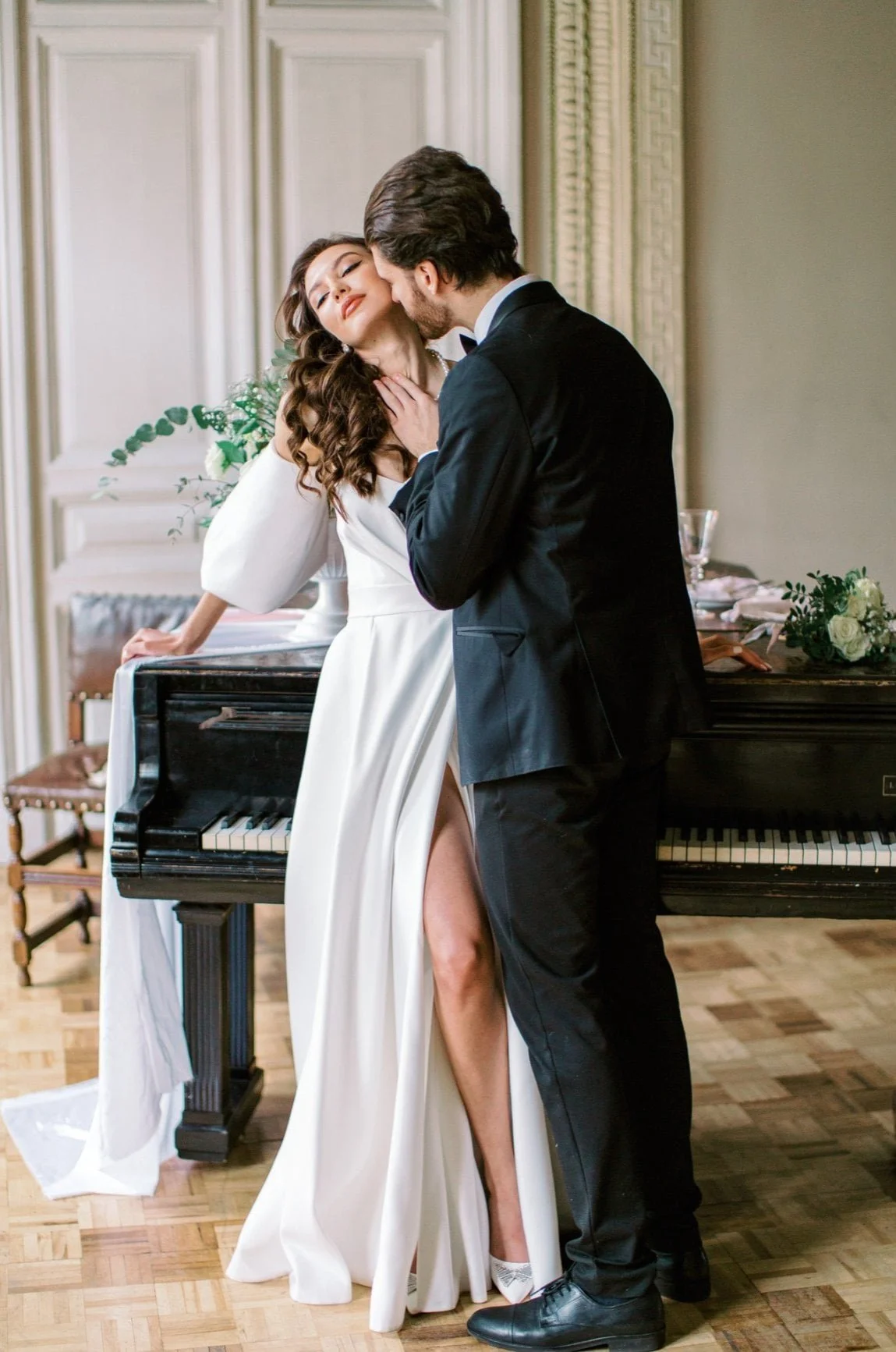 A couple dressed in formal attire on a dance floor, with the woman leaning against a piano and the man holding her close, in an elegant room with ornate moldings and a floral arrangement on the piano.