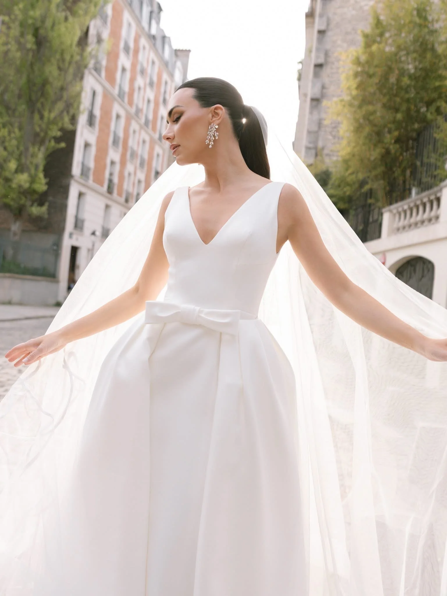 A bride in a white wedding dress with a V-neckline and bow detail, standing outdoors between tall buildings and trees, with sunlight behind her and a long veil flowing behind.