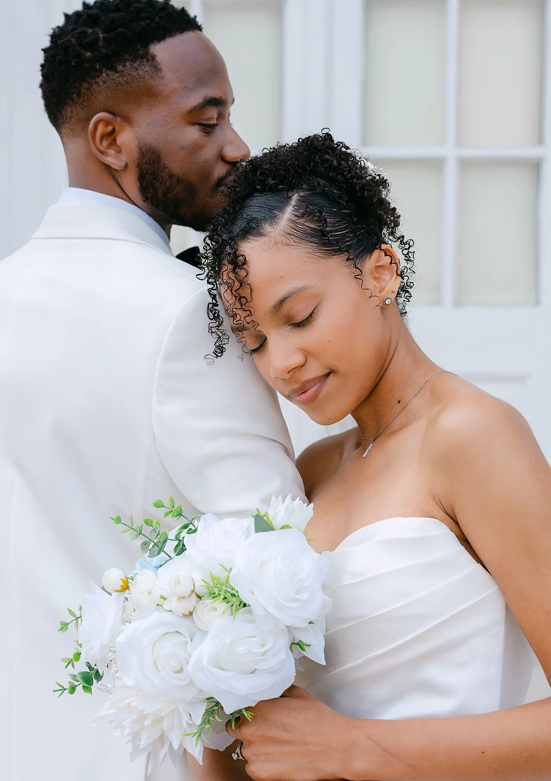 A bride and groom embracing on their wedding day, with the bride holding a bouquet of white flowers and the groom in a white suit. They are smiling with closed eyes, showing affection and happiness.