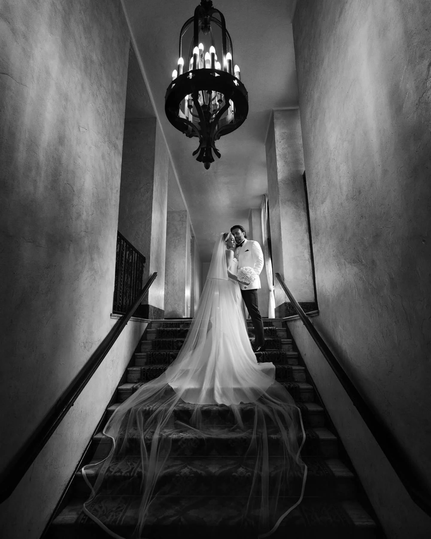Black and white photo of a bride and groom standing on a staircase, smiling and embracing, with a chandelier above them, and she is wearing a long wedding dress with a veil, he is wearing a tuxedo.