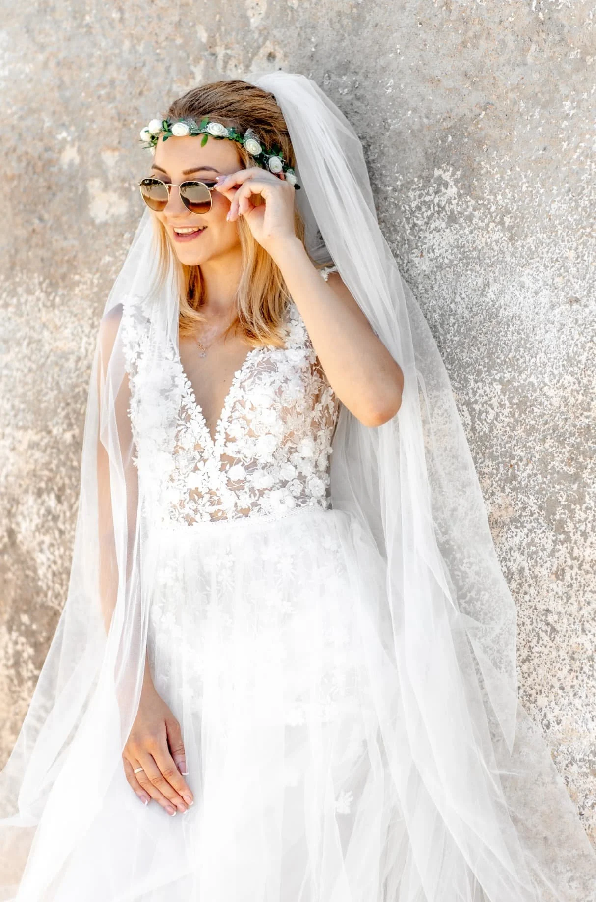 A woman wearing a wedding dress with lace and floral details, a veil, sunglasses, and a floral headband, standing against a textured wall.