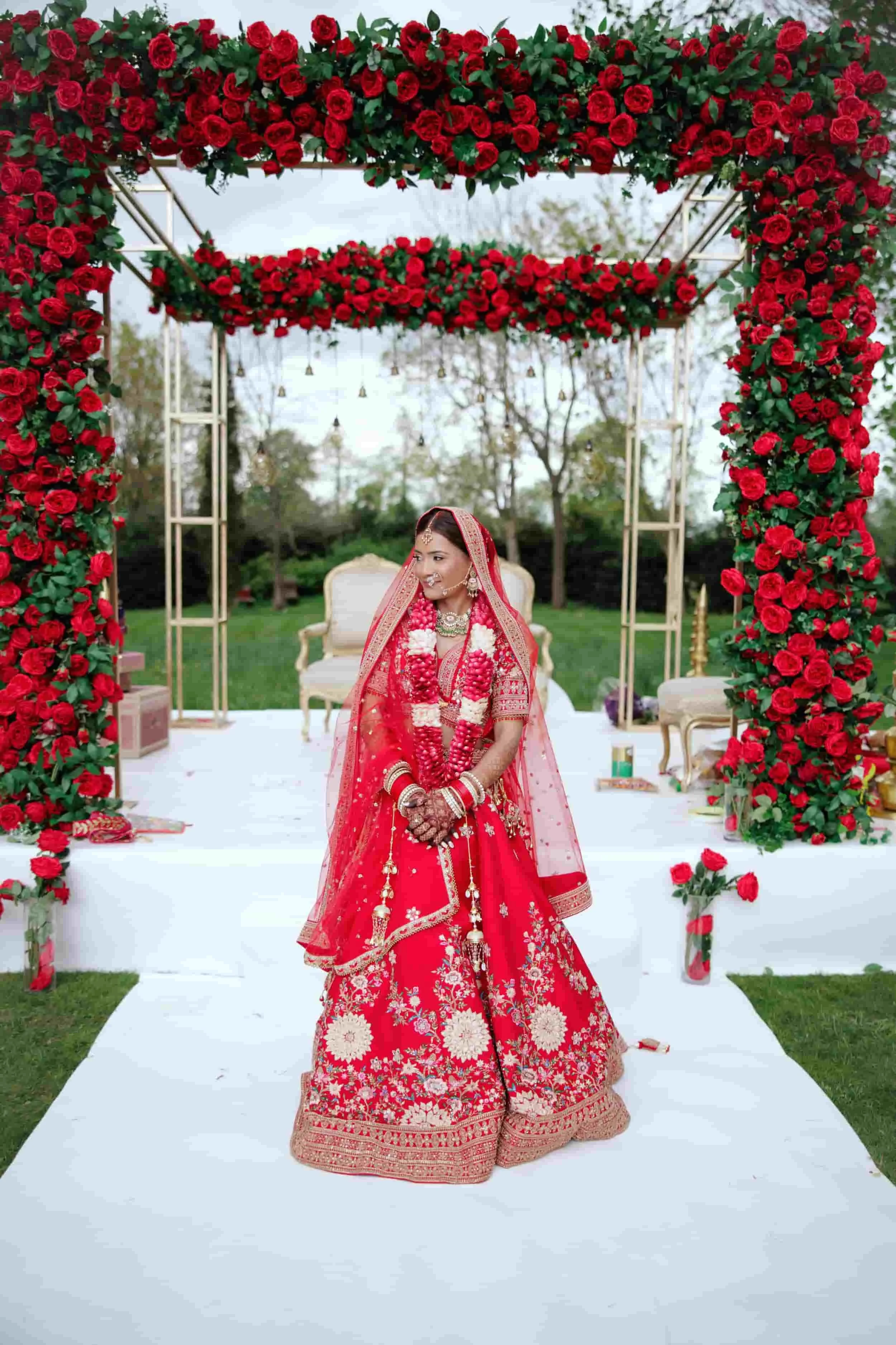 A bride in a red traditional Indian wedding dress with gold embroidery, standing under a floral arch made of red roses, smiling. The ceremony is outdoors with trees in the background.
