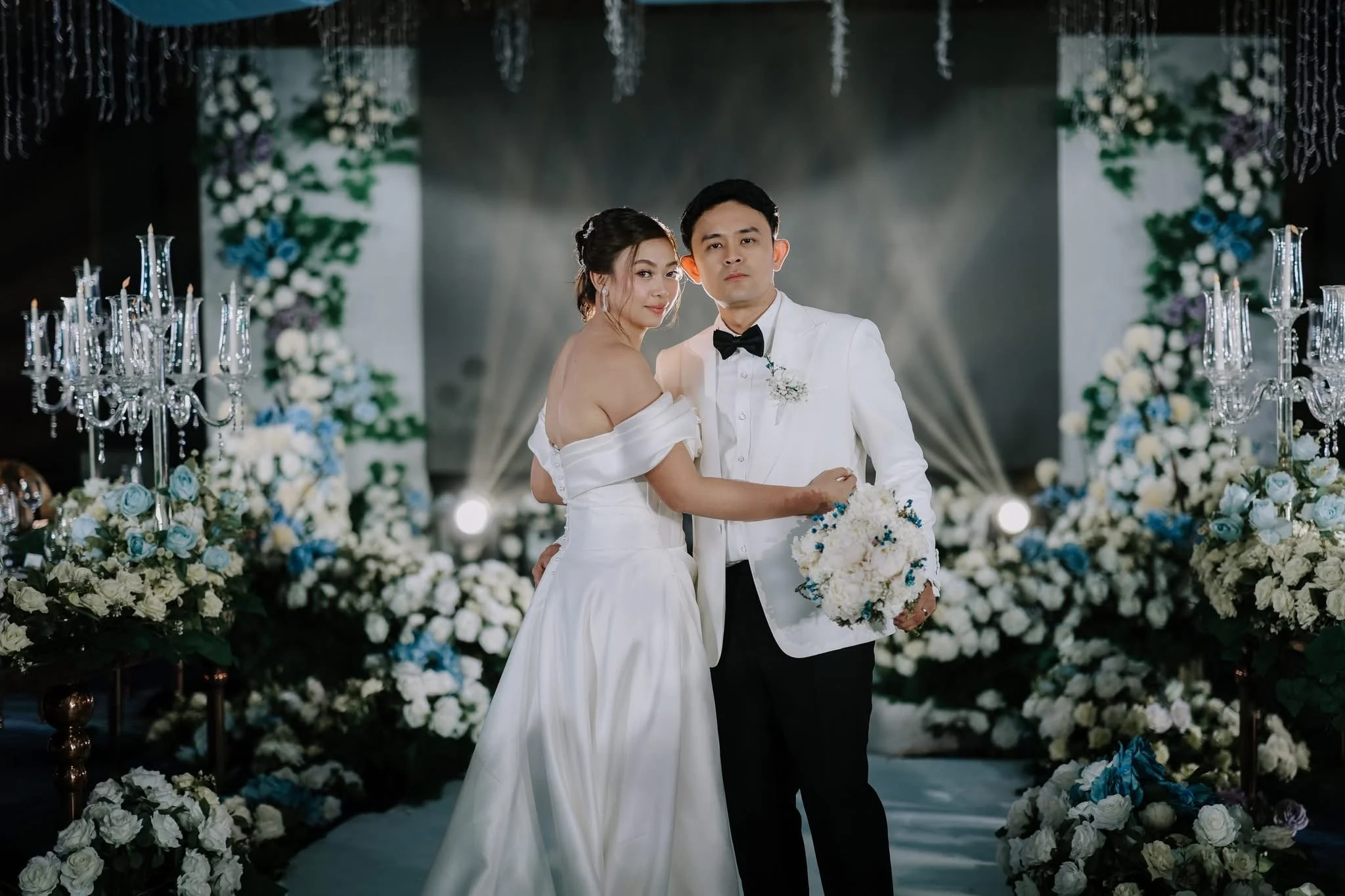 A bride and groom in wedding attire standing close together amid a decorative floral setting with white, blue, and purple flowers and candelabras.