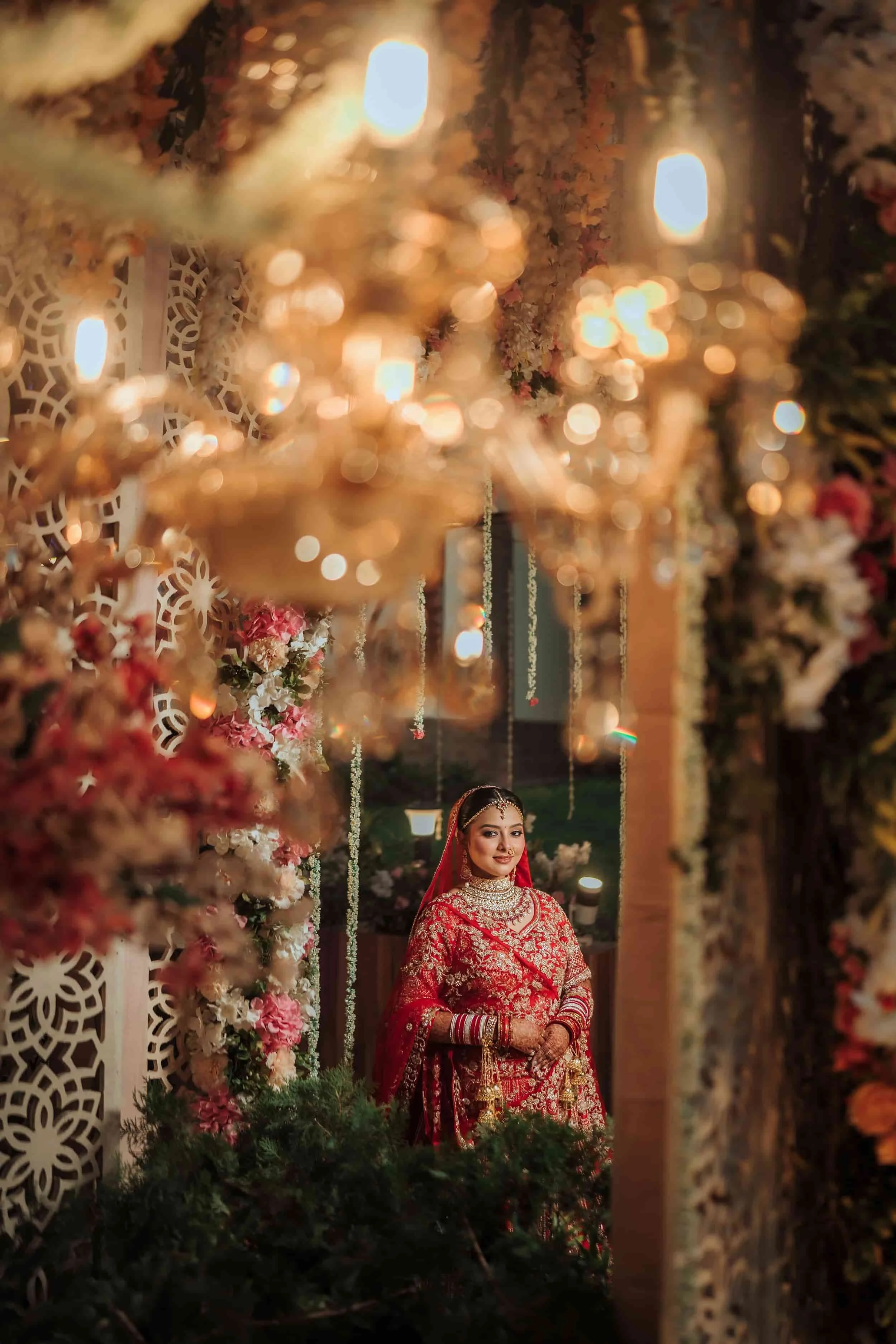A young woman dressed in a traditional red and gold bridal outfit, standing in a decorated venue with floral and ornate decor, illuminated by warm lights.