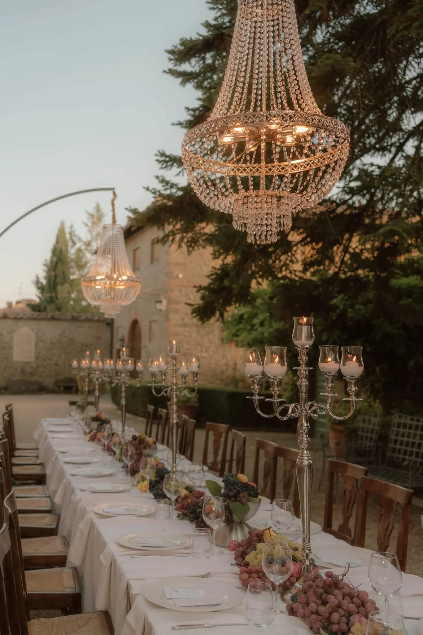 An outdoor dining area decorated for an event with a long table, elegant chandeliers hanging above, and tall candelabra with lit candles. The table is set with white tablecloths, napkins, glassware, plates, and floral centerpieces with grapes and oth