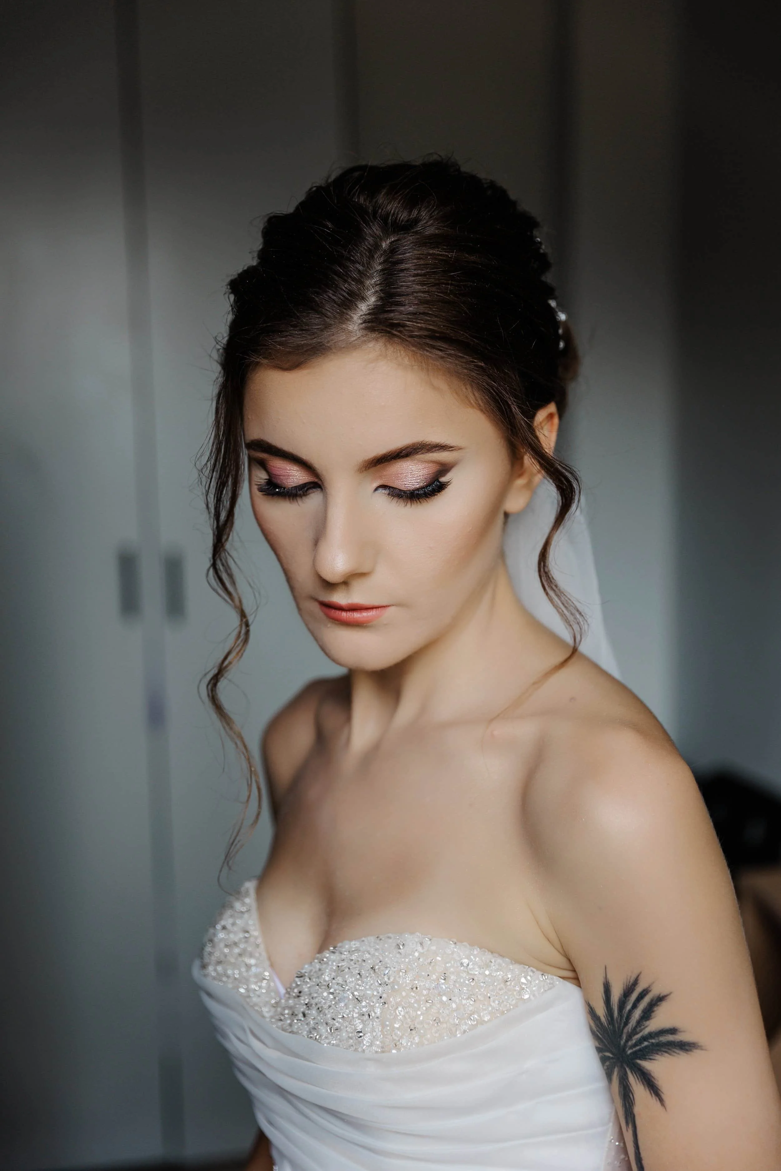 Close-up of a bride with styled hair, makeup, wearing a white wedding dress with beaded details, and a palm tree tattoo on her arm.