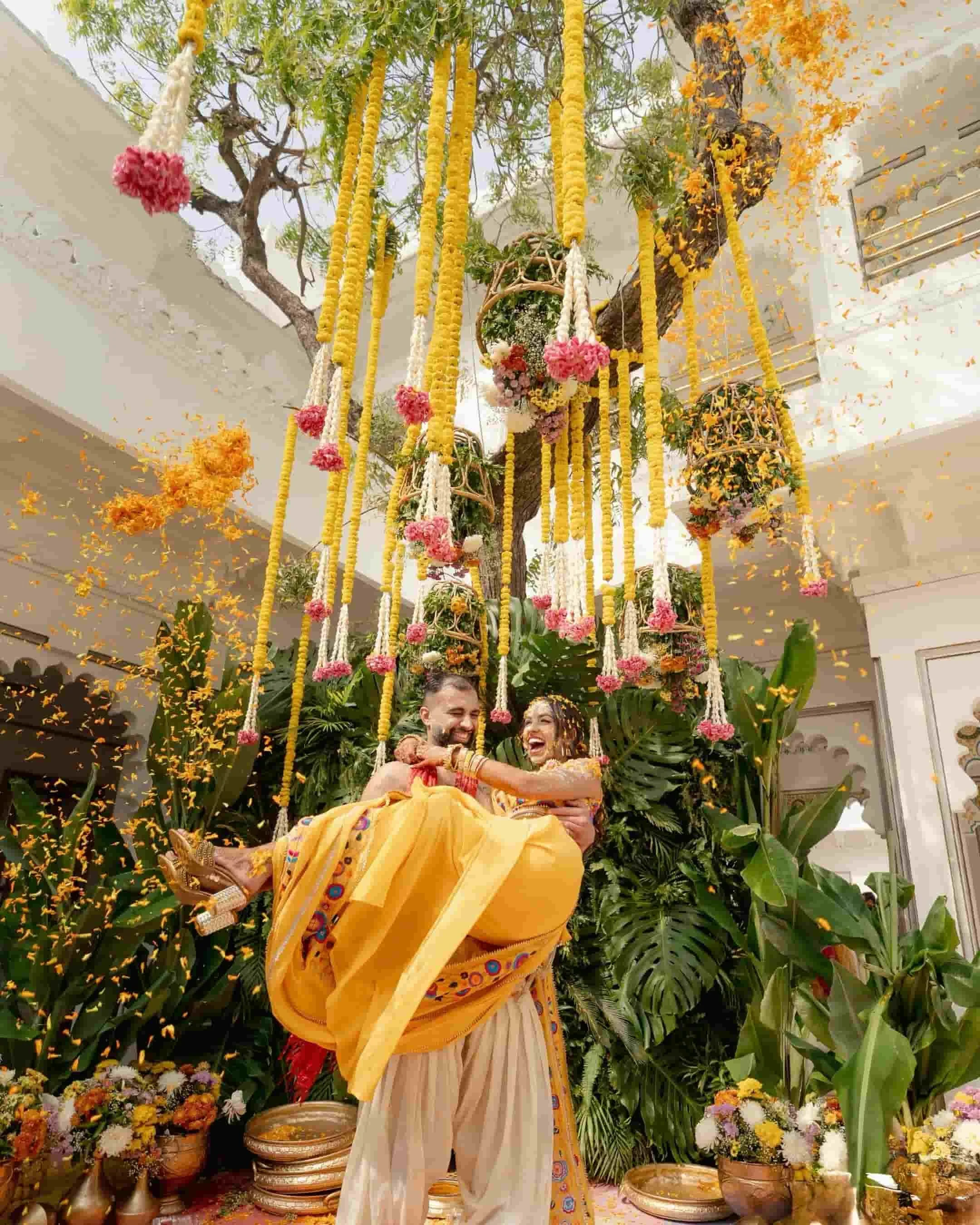 A traditional Indian wedding ceremony with a bride and groom sitting on a swing decorated with yellow marigold flowers, surrounded by lush green plants and vibrant flowers, during the daytime.