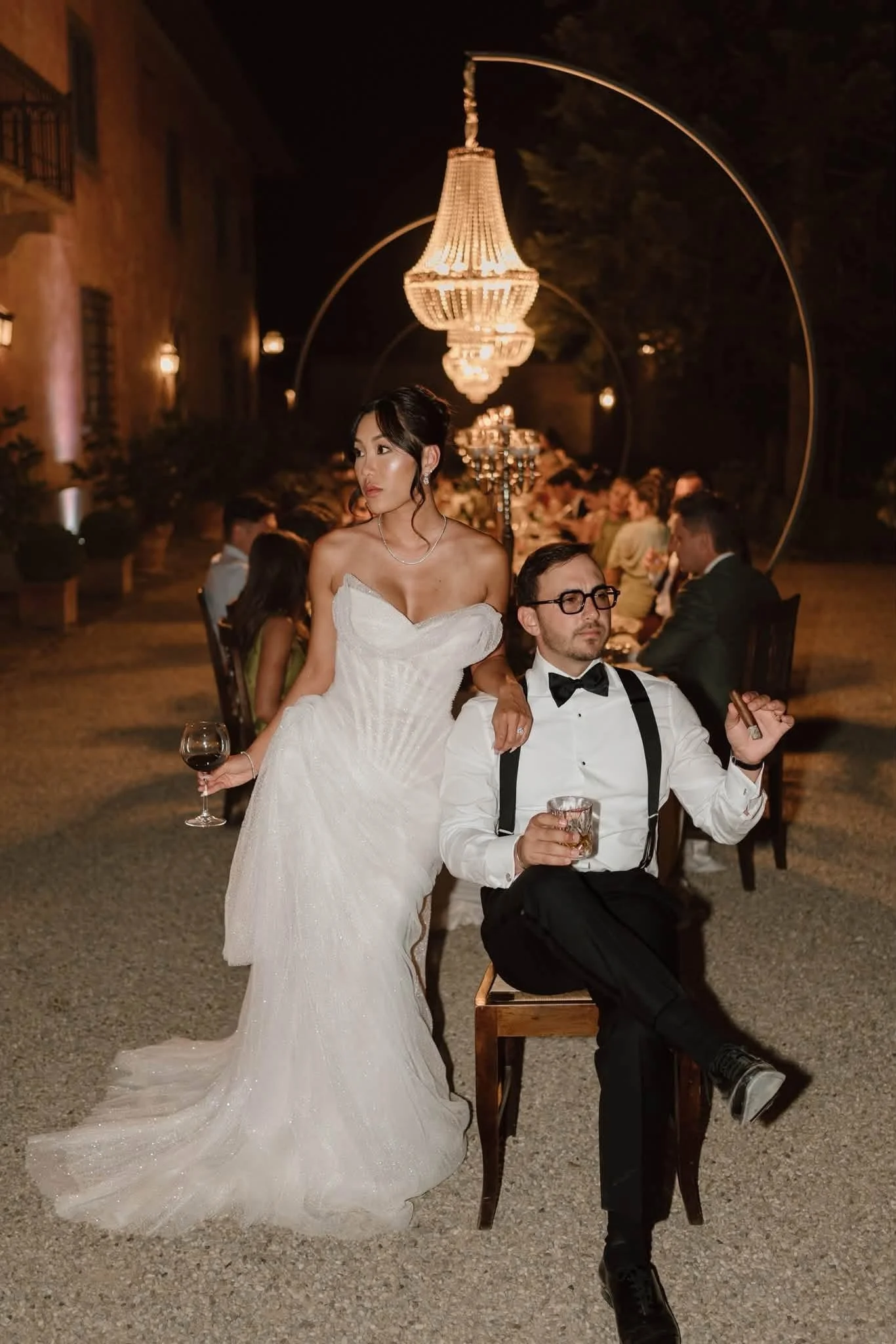 A bride in a white wedding dress holding a glass of red wine, standing next to a seated groom with glasses, in an outdoor evening setting with a large chandelier and guests in the background.