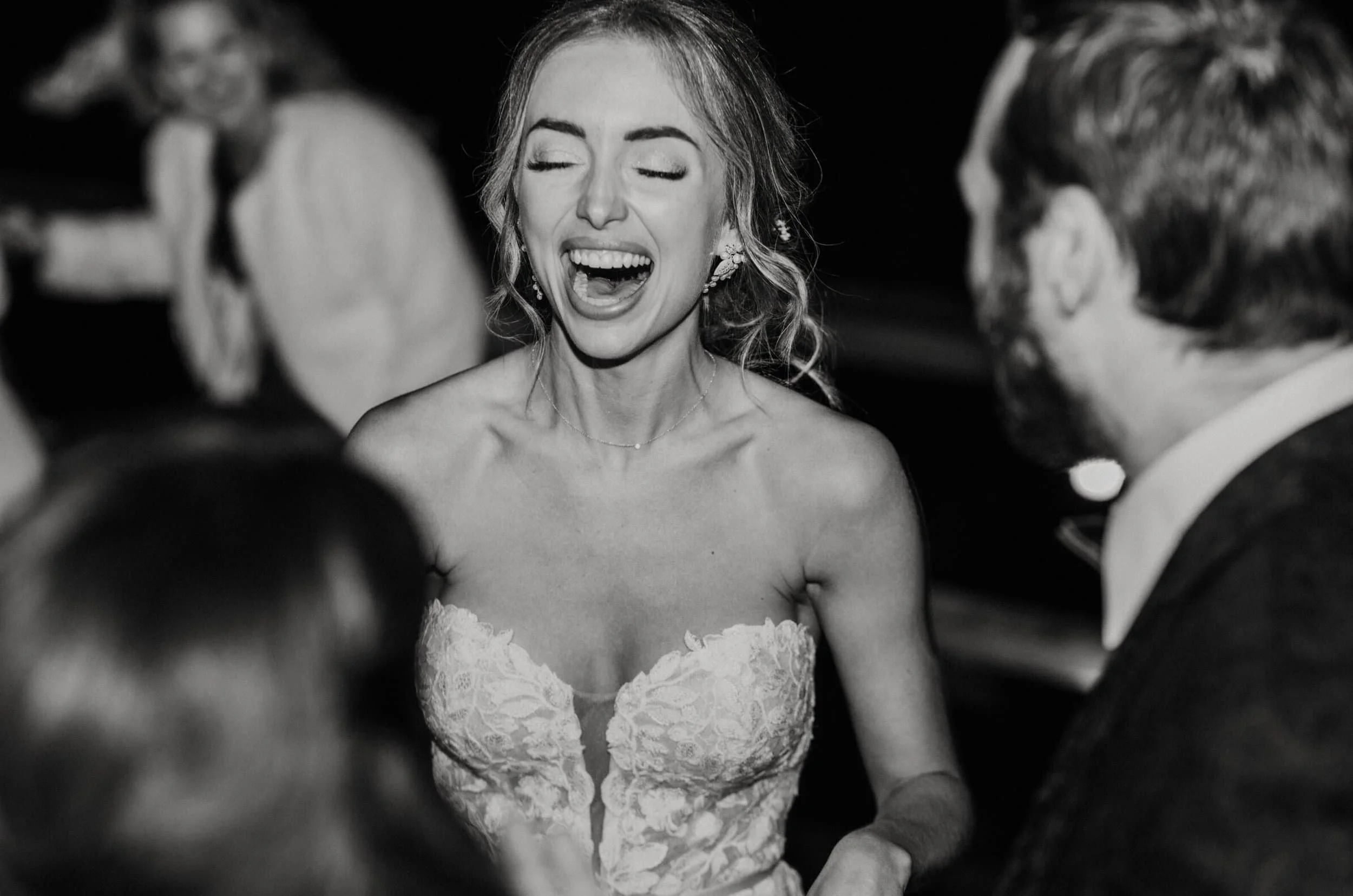 Black and white photo of a woman with her eyes closed, smiling widely, surrounded by a group of people, likely at a formal event.