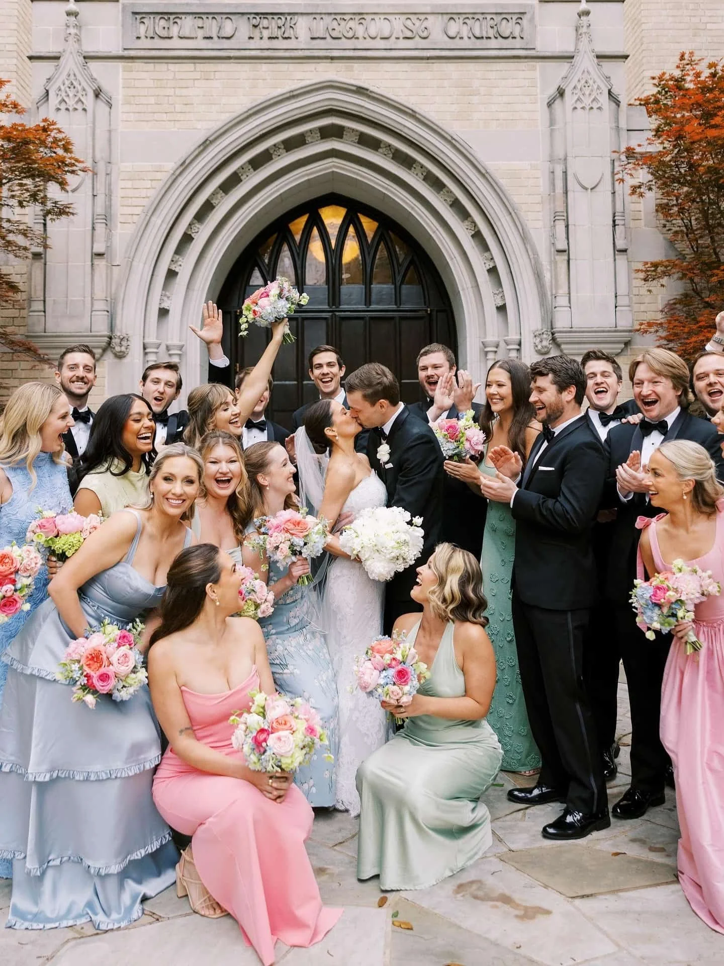 A large wedding party outside a church, with a bride and groom kissing in the center surrounded by bridesmaids holding bouquets of pink flowers and groomsmen in tuxedos, celebrating after their wedding.