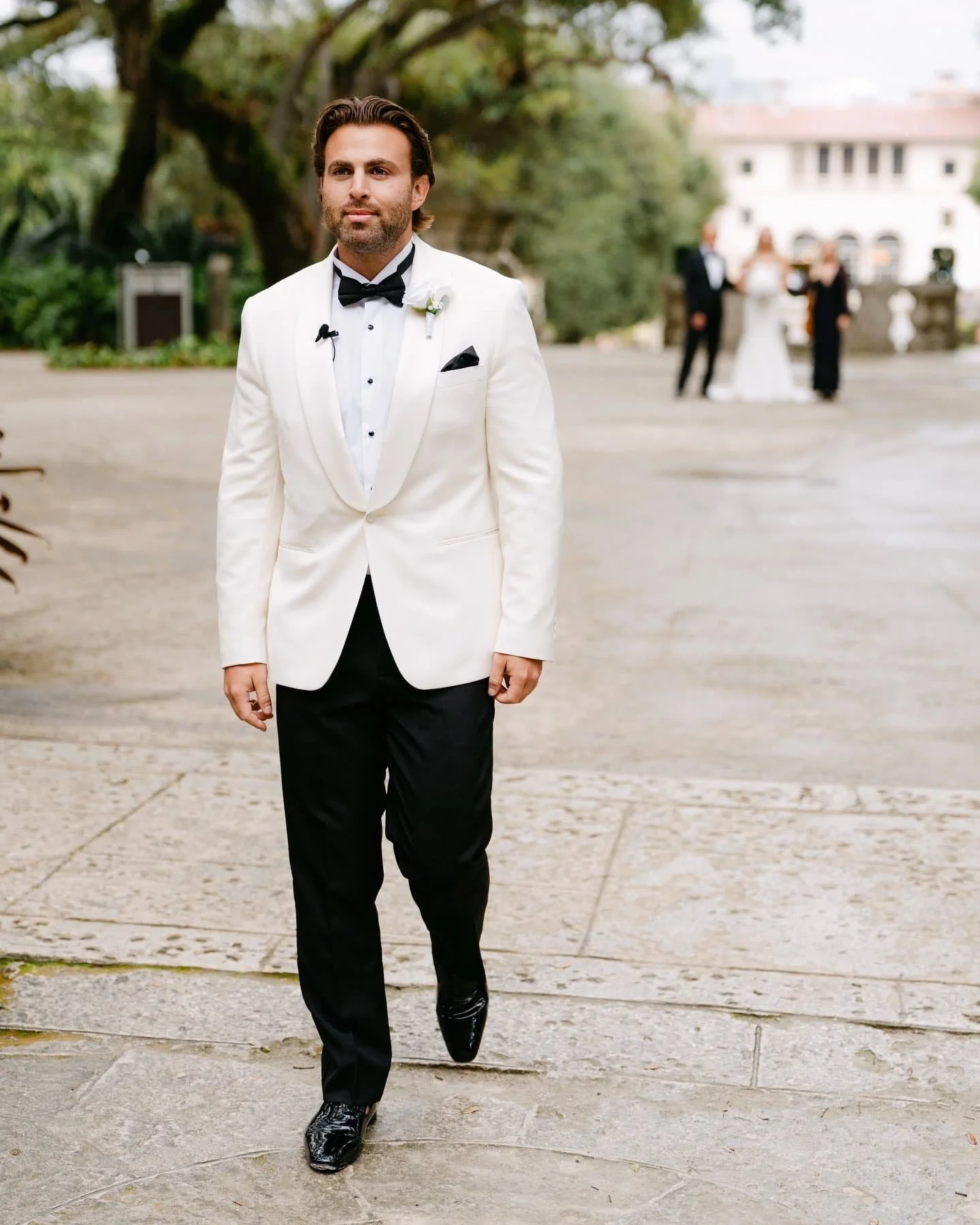 A man in a white tuxedo and black bow tie walking outdoors, with a blurred group of people in formal attire in the background.