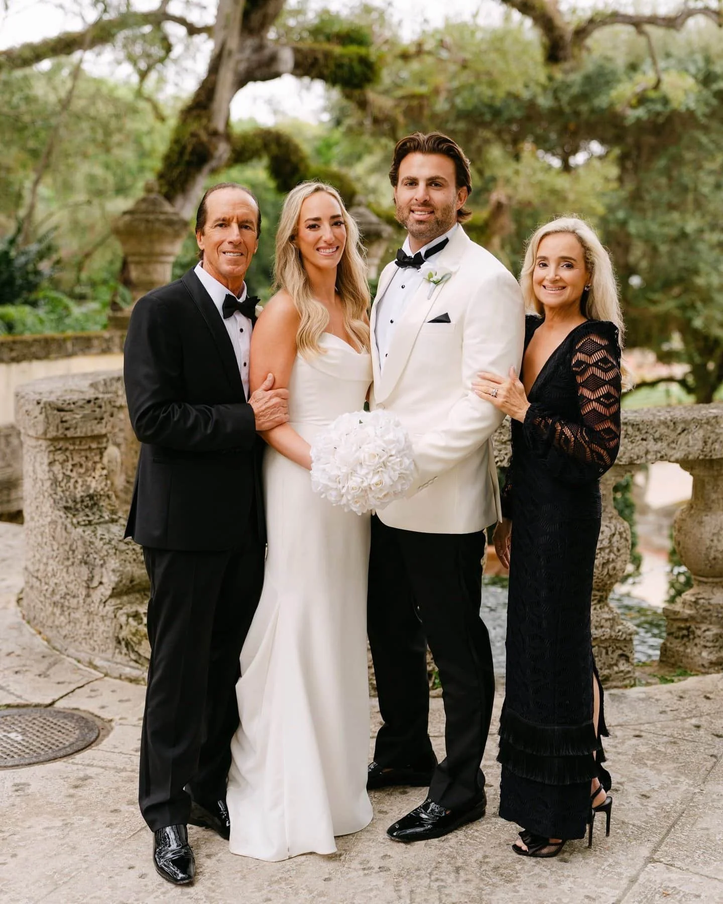Group of five people at a wedding, including groom, bride, and family, standing outdoors near an ornate stone fountain with trees in the background.