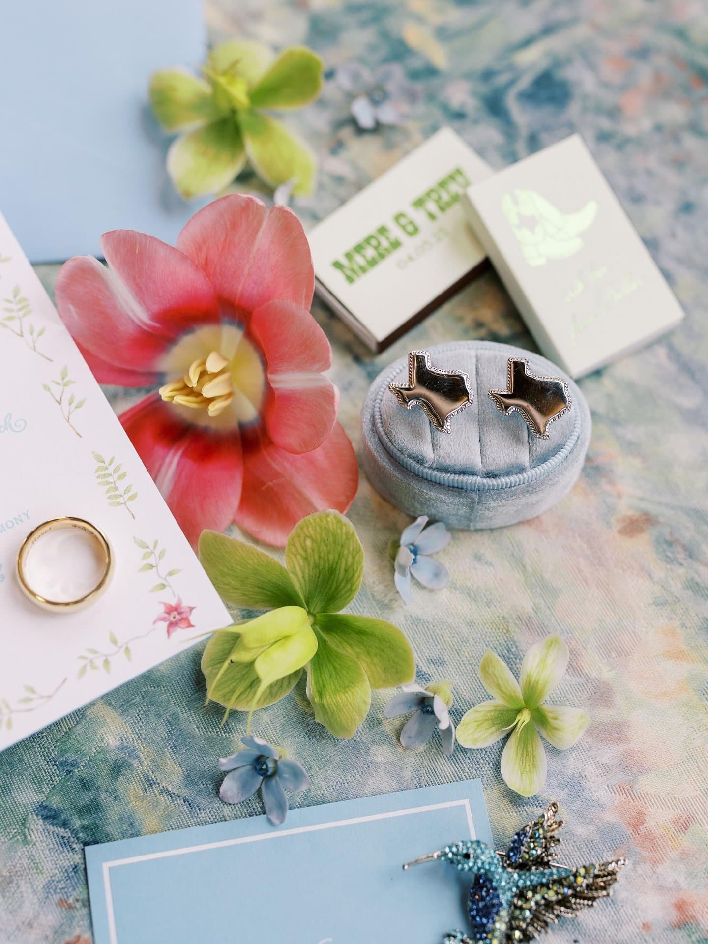 Close-up of jewelry collection with a gold wedding band, blue dragonfly brooch, and decorative boxes, surrounded by pastel-colored flowers and leaves on a floral fabric surface.