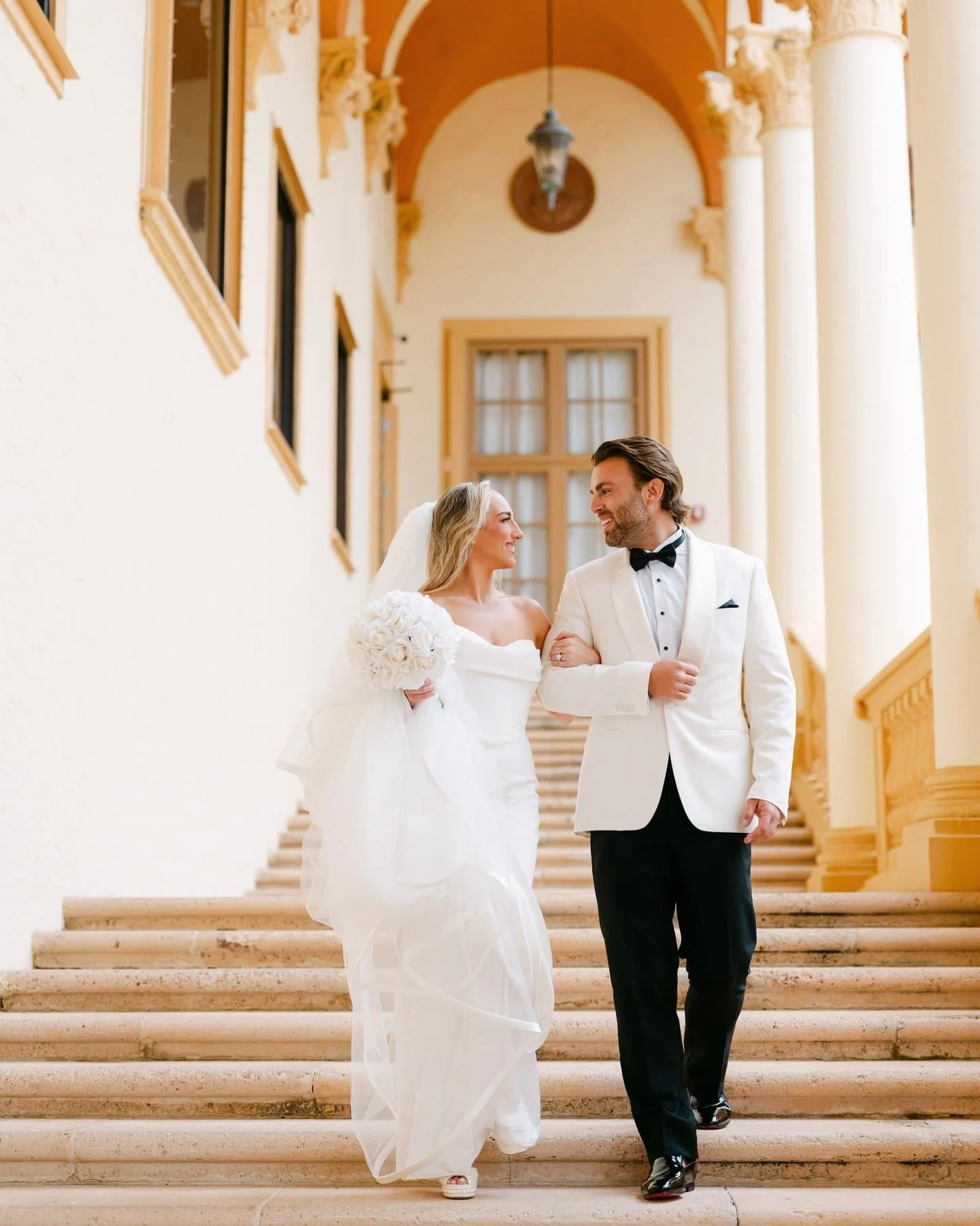 A bride and groom walking down the stairs of a grand, elegant building with columns. The bride is wearing a white wedding gown, holding a bouquet of white flowers, and the groom is dressed in a white tuxedo jacket with a black bow tie and black trous
