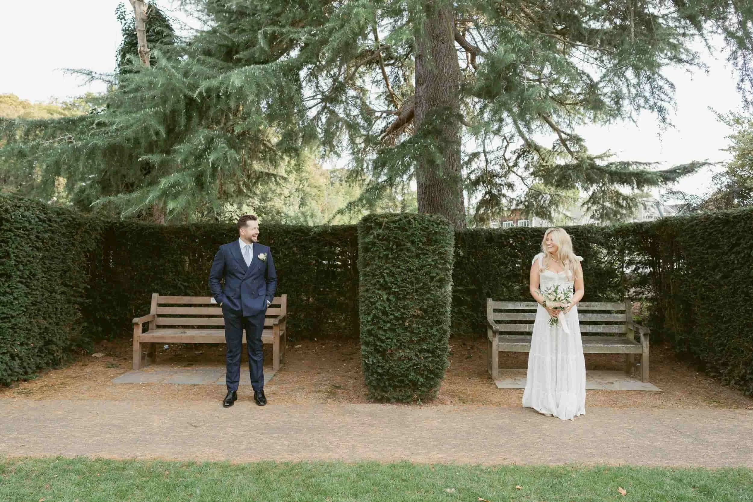 A man and a woman dressed in wedding attire standing outdoors on a pathway in front of a large evergreen tree surrounded by a hedge. The man is wearing a blue suit and tie, smiling, looking at the woman. The woman is wearing a long white wedding dres