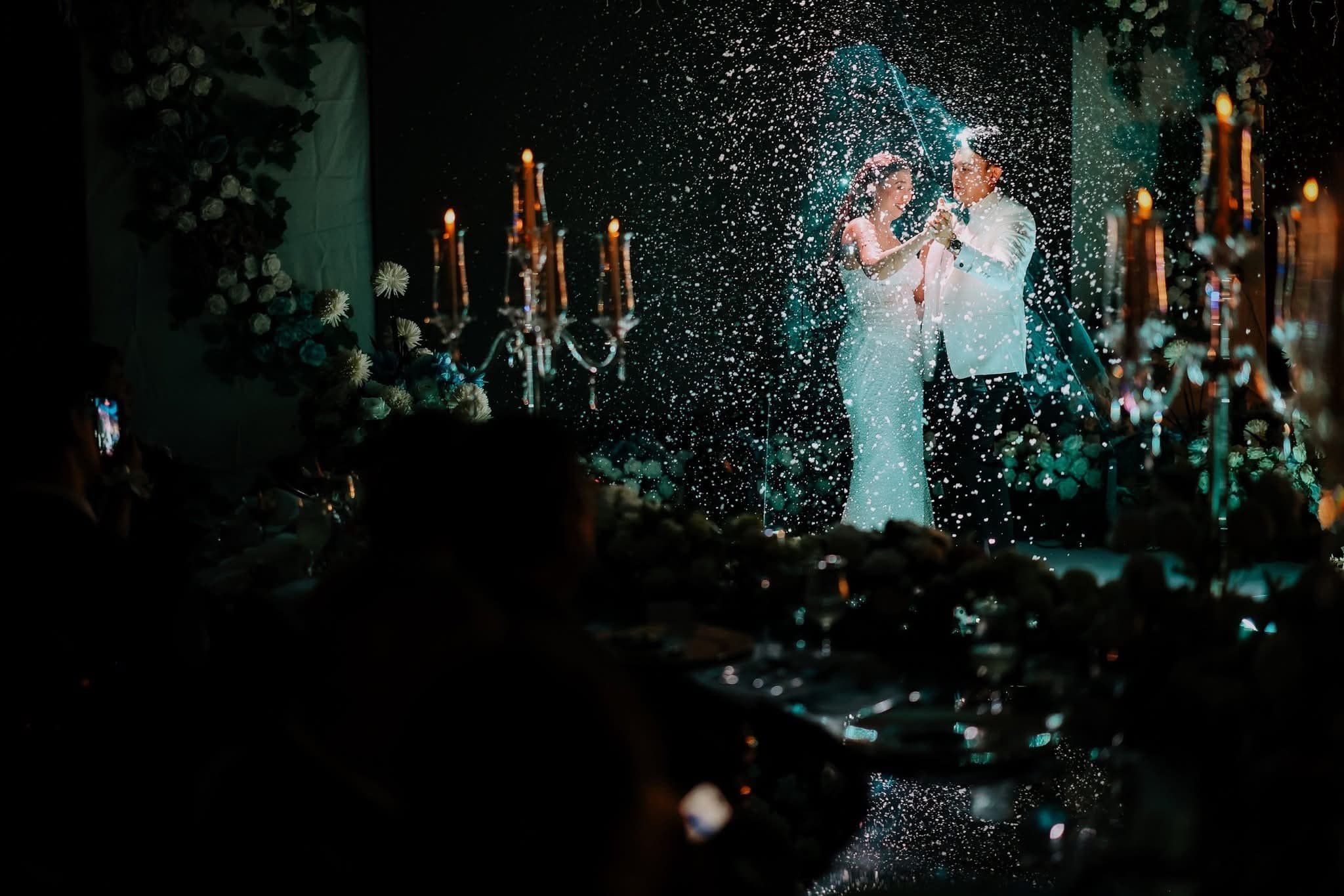 A couple dancing at their wedding reception, illuminated by candlelight, with a sparkler or firework display creating a shower of sparks around them.