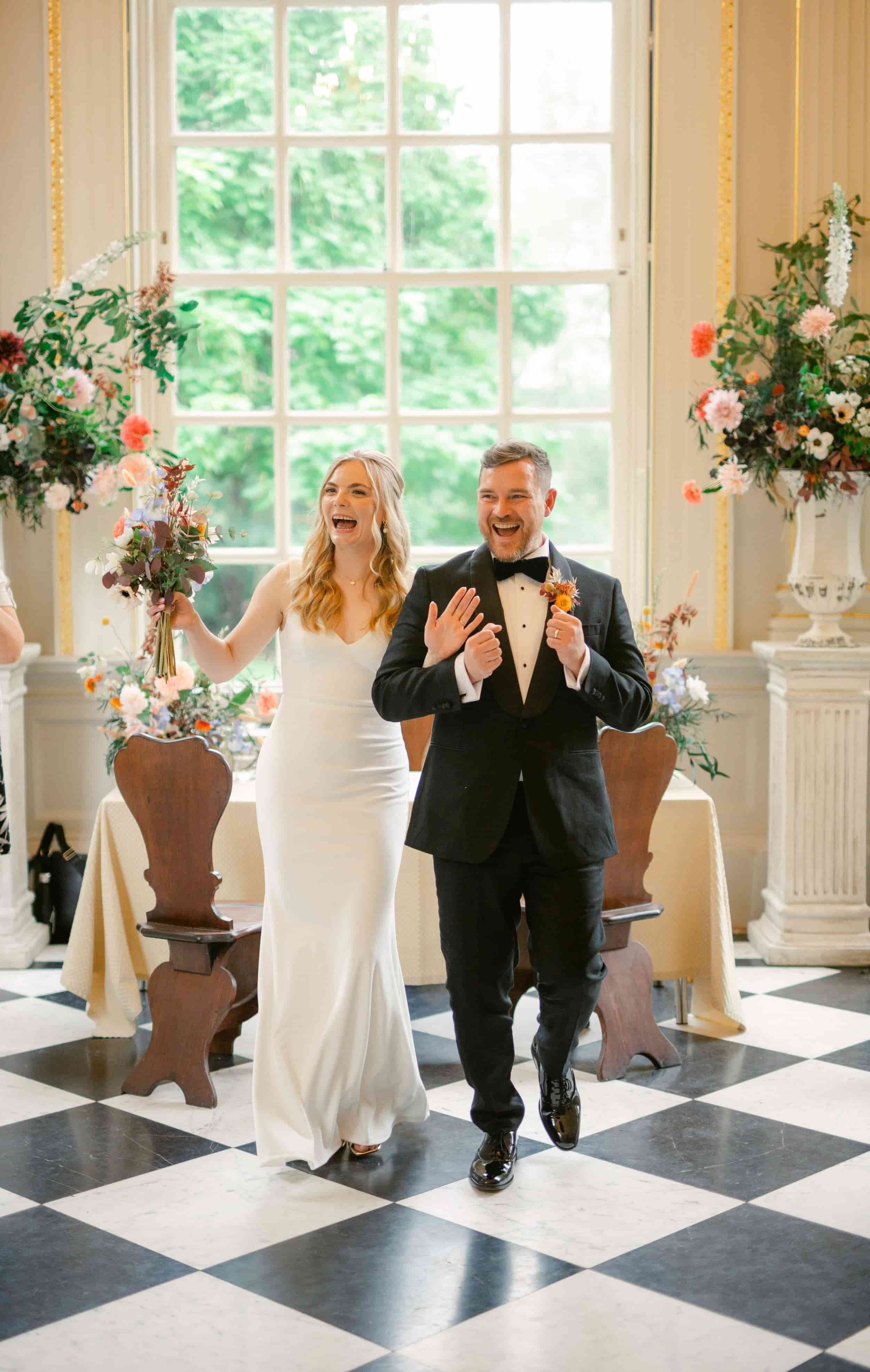 A bride and groom walking into a wedding venue, smiling and joyful, surrounded by floral arrangements, with a large window and greenery outside.