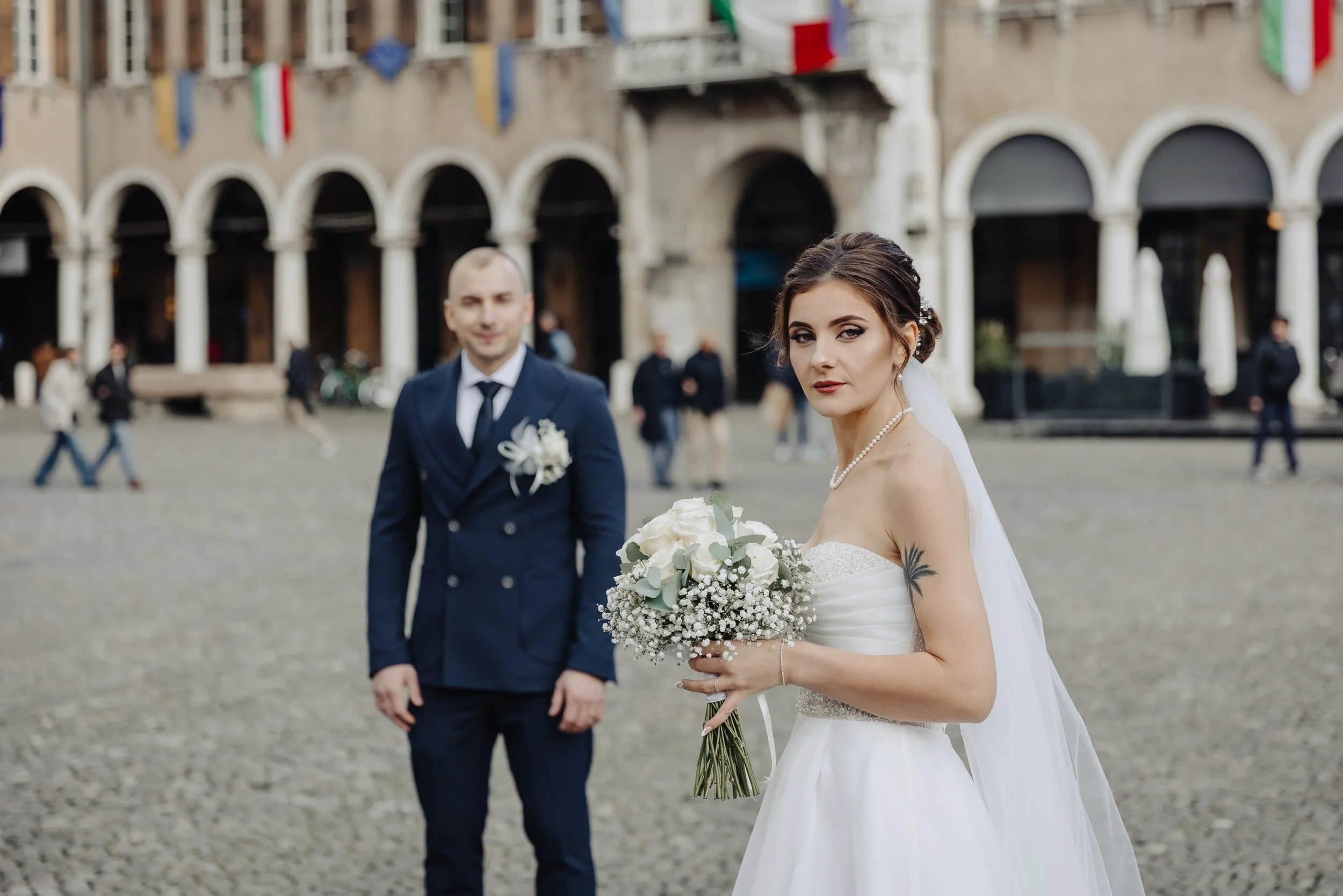 A bride holding a bouquet of white roses and baby's breath stands outdoors in front of a building with arched windows and flags hanging outside. A groom in a blue suit with a boutonniere is blurred in the background.