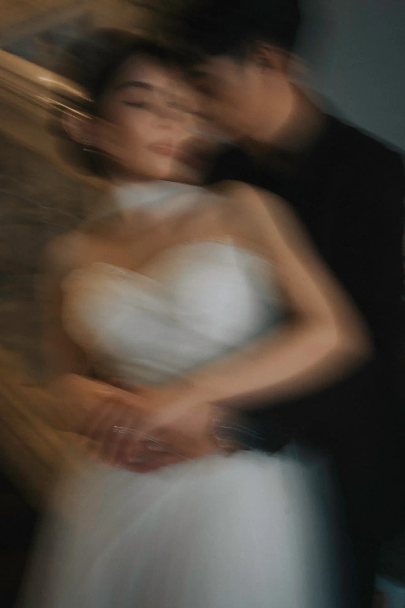 A blurry photo of a person with dark hair, wearing a white top, holding their face with their hand, and lying down on a surface with a dark background.