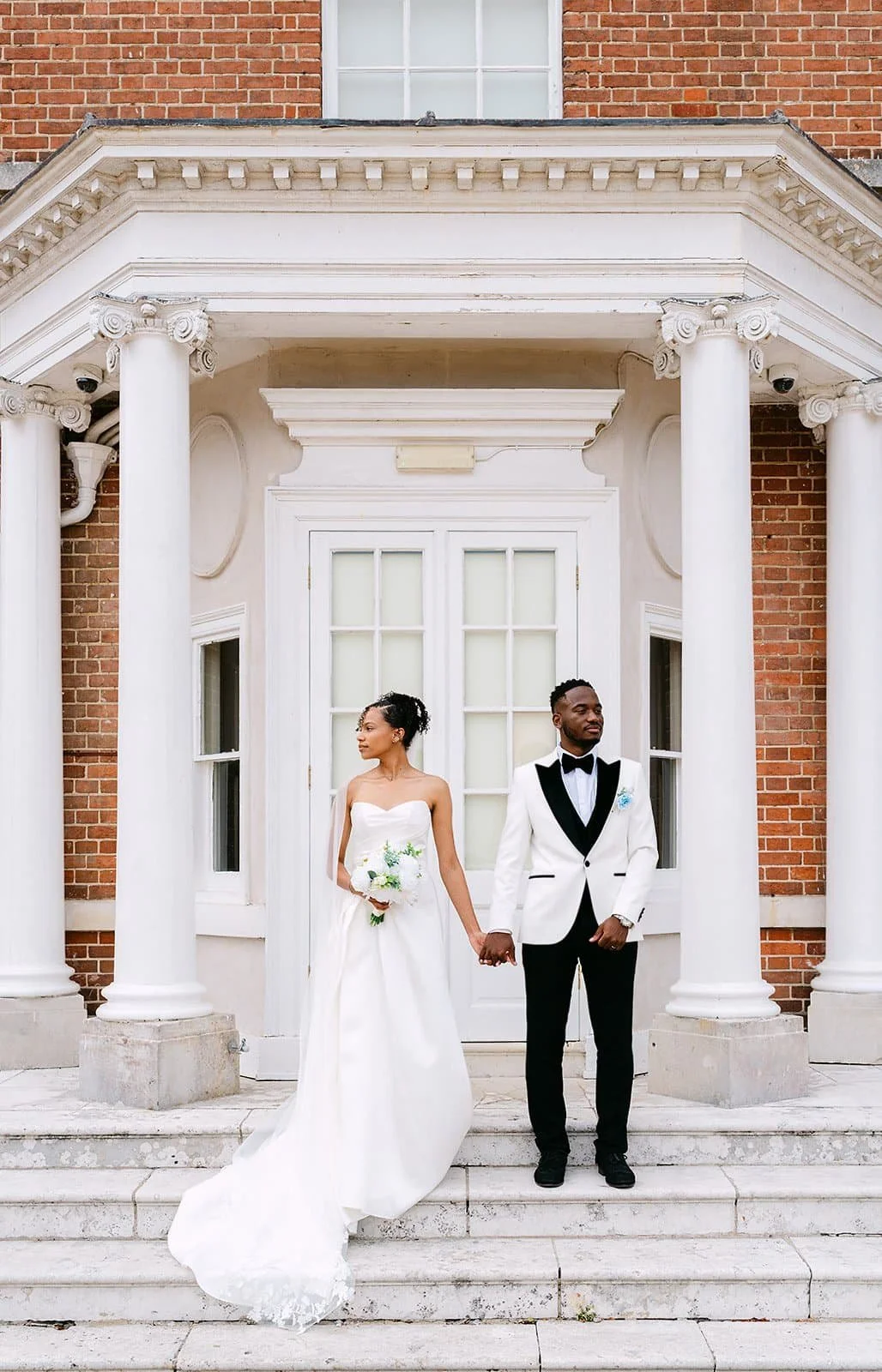 A bride and groom holding hands on the steps of a brick building with white columns and large window behind them. The bride in a strapless white wedding gown, holding a bouquet, and the groom in a tuxedo with black lapels and bow tie.