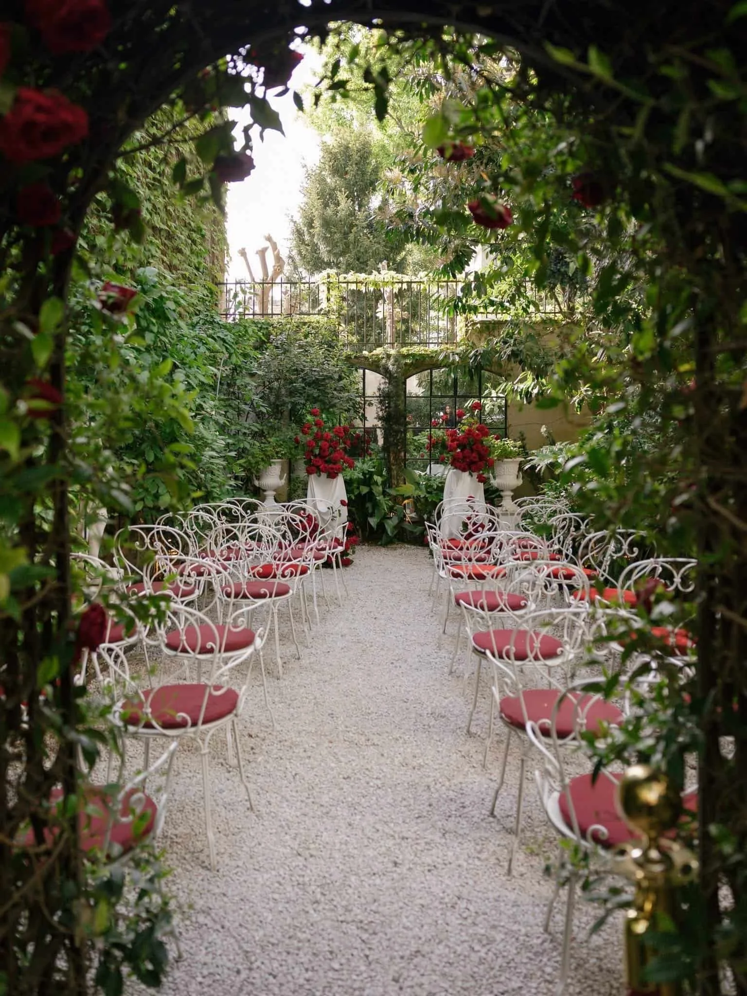 A quaint outdoor wedding venue surrounded by green foliage, with white wrought iron chairs with red cushions arranged on a white gravel aisle, red floral arrangements in white urns, and a black metal arch decorated with red flowers at the end of the 
