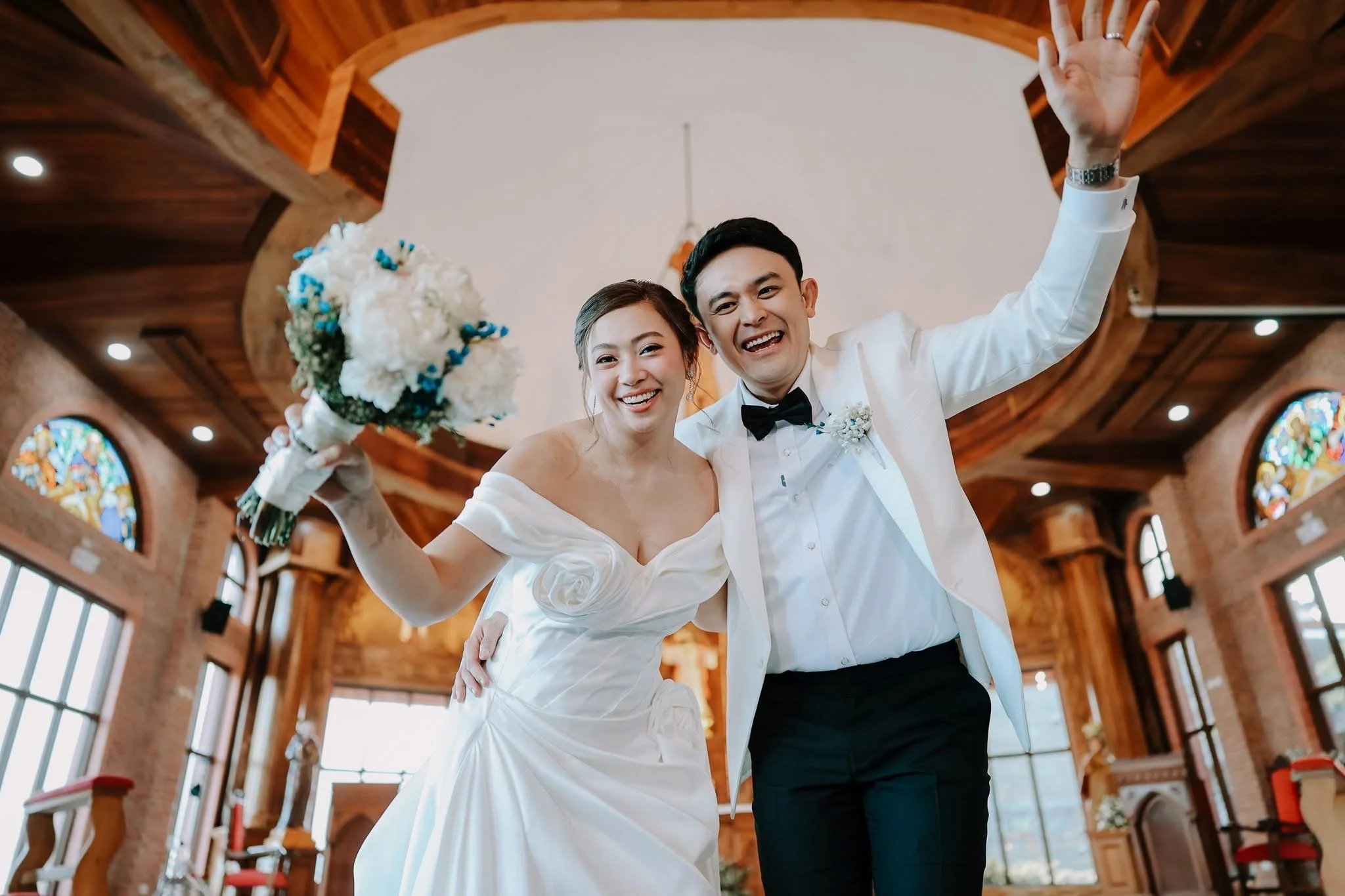 A happy bride and groom smiling and waving at the camera inside a wooden church, with stained glass windows in the background.