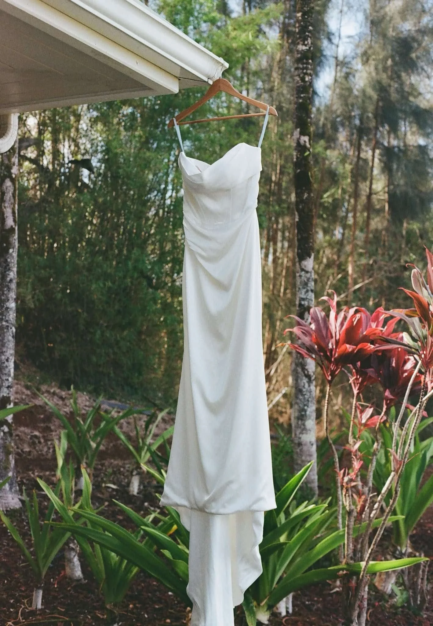 A white fabric dress draped over a wooden hanger, hanging from the edge of a house roof, with trees and plants in the background.
