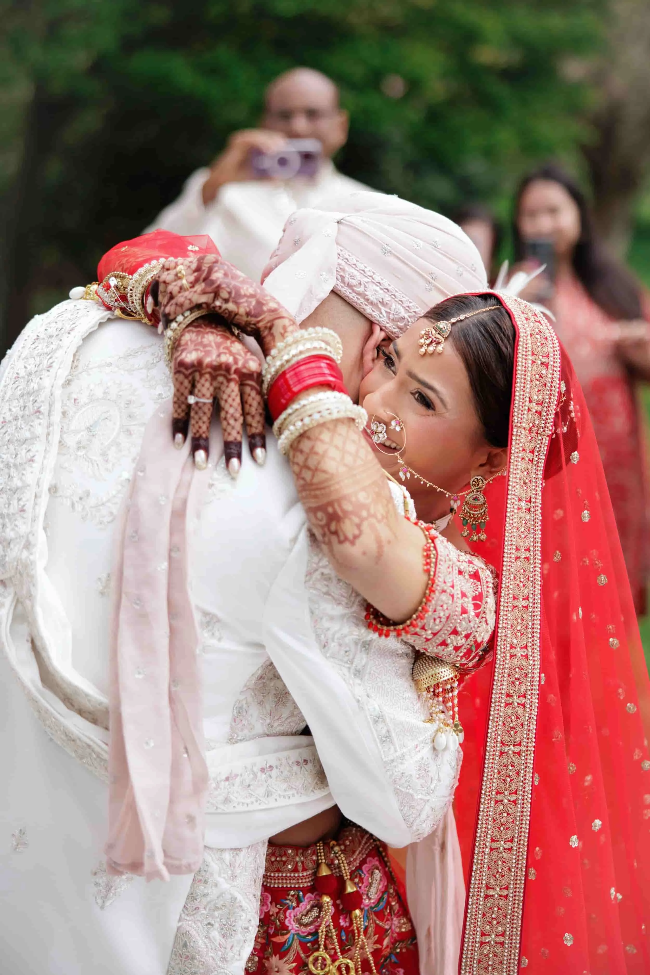 A bride dressed in red and gold traditional Indian attire hugs a person dressed in white at a wedding ceremony, with guests in the background taking photos.