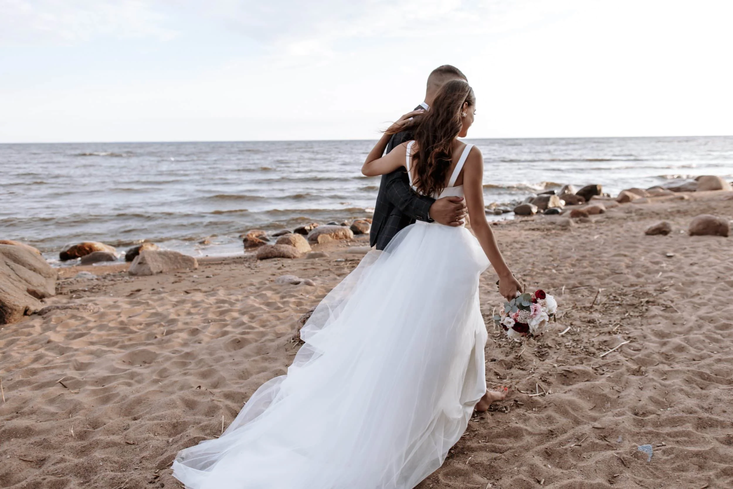 Couple in wedding attire walking arm in arm along a sandy beach with rocks, with the ocean and cloudy sky in the background.