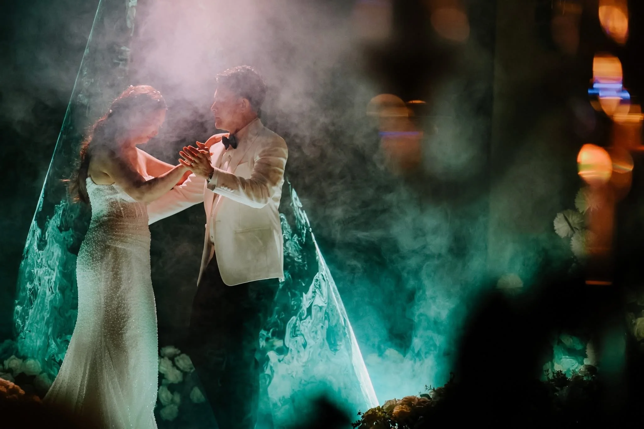 A bride and groom hold hands while dancing in a dreamy, misty setting with soft lighting and floral decorations.