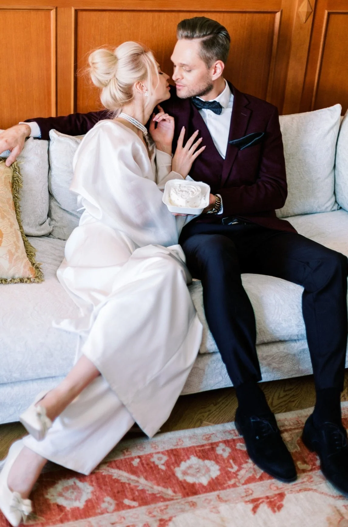 A man and a woman are sitting on a sofa in a cozy room with wooden walls. The woman, dressed in a white satin robe, is holding a container of ice cream and leaning in close to the man. The man, wearing a dark suit with a bow tie, is touching her face