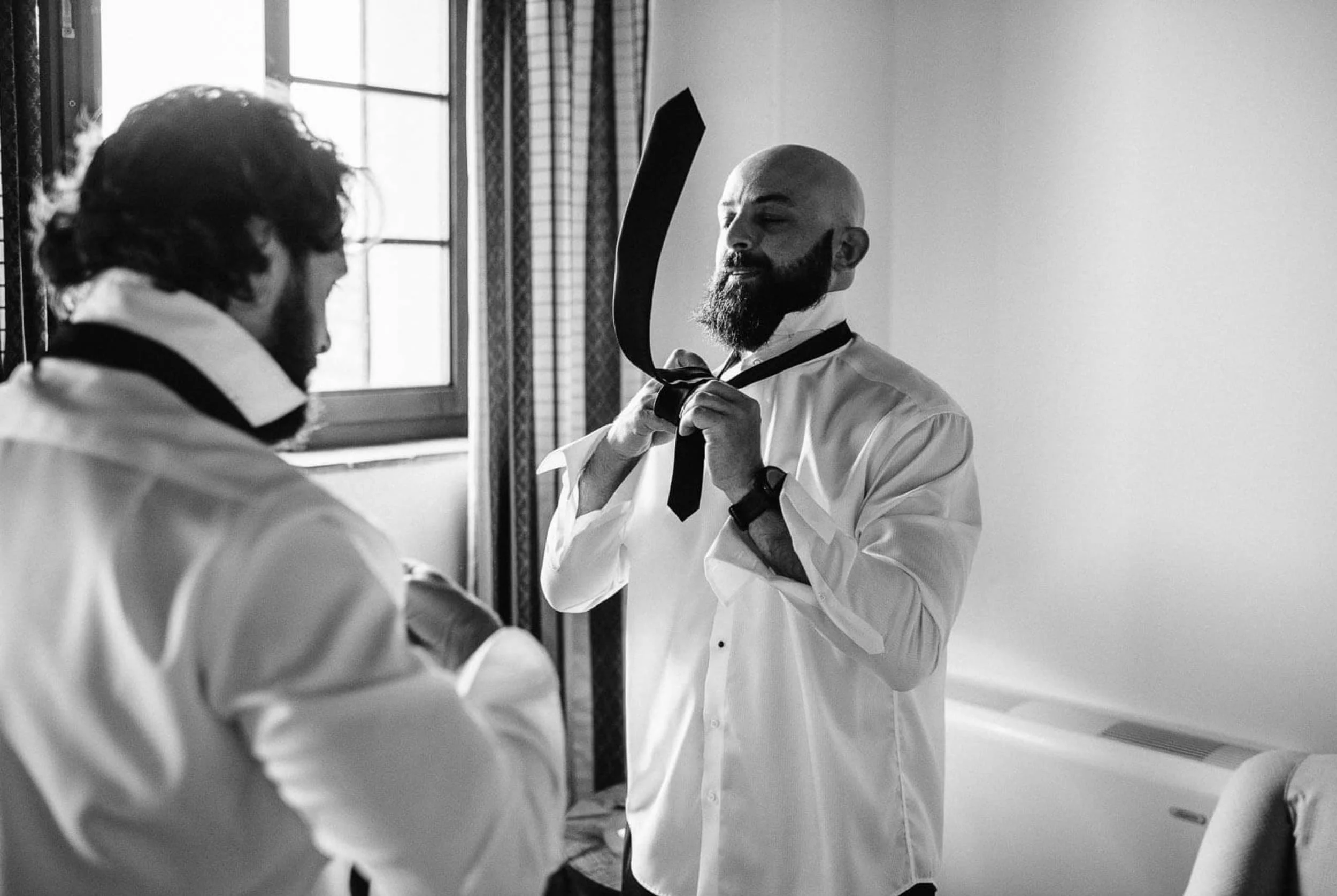 A man with a beard adjusting a necktie in front of a mirror, with another person partially visible in the foreground. The scene is lit by natural light from a window.