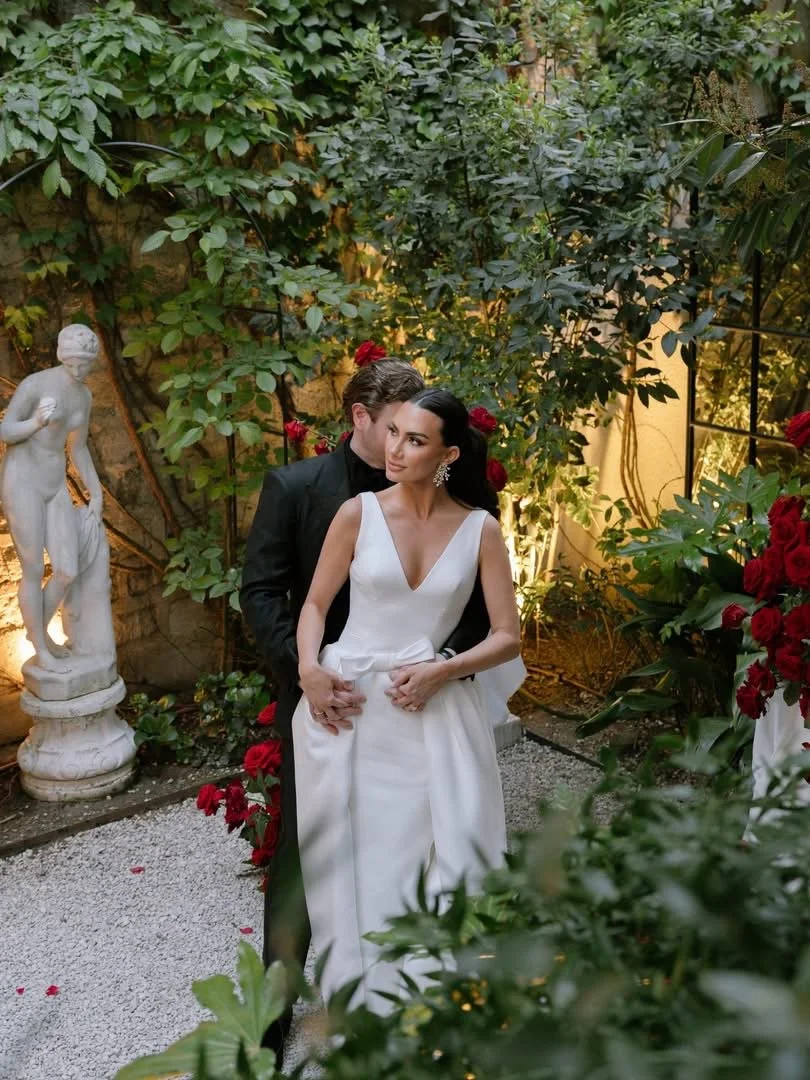 A bride and groom standing together in an outdoor garden setting surrounded by green foliage and red roses, with a classical statue in the background.