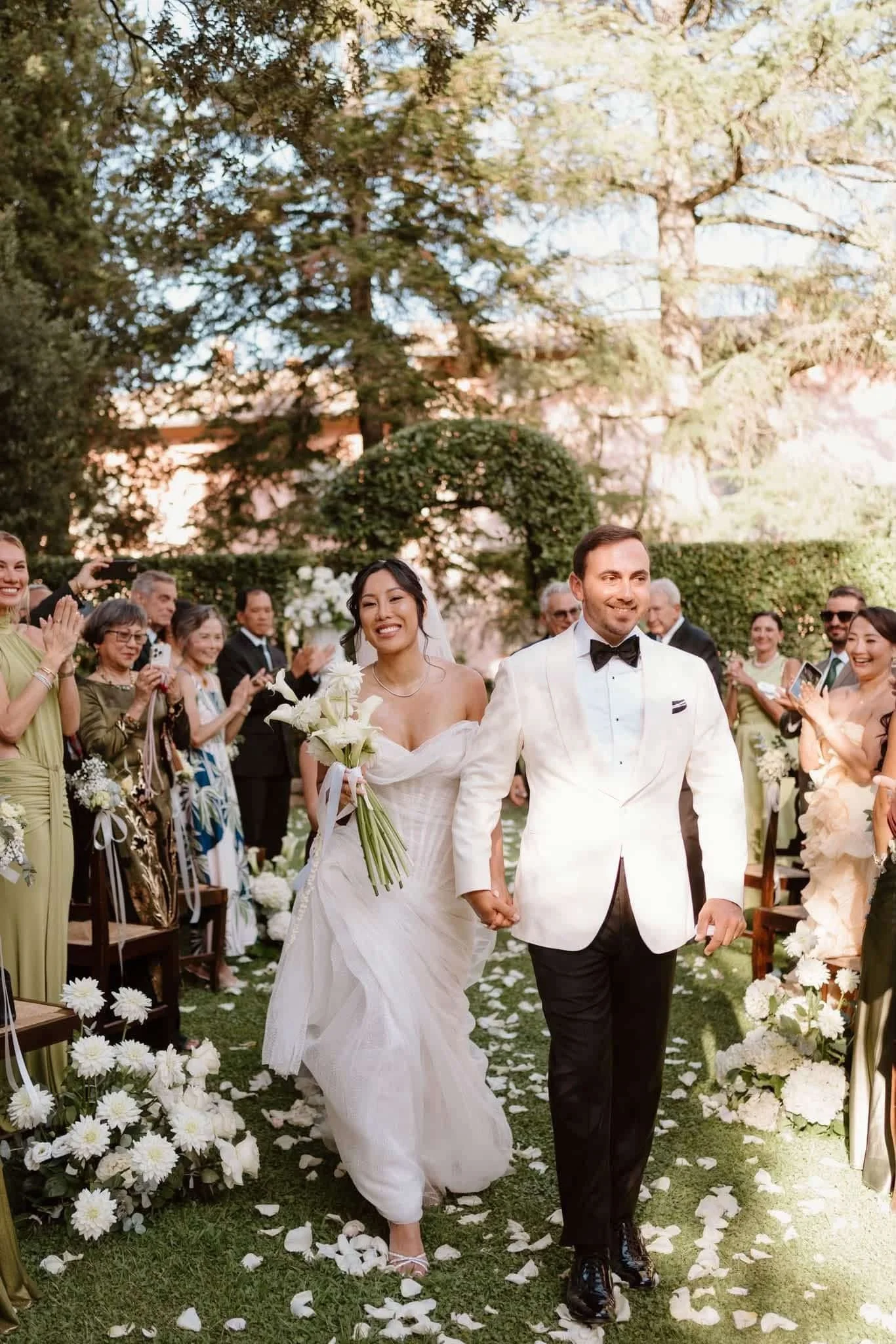 A bride and groom walking hand in hand down the aisle of an outdoor wedding ceremony, surrounded by guests and floral decorations.
