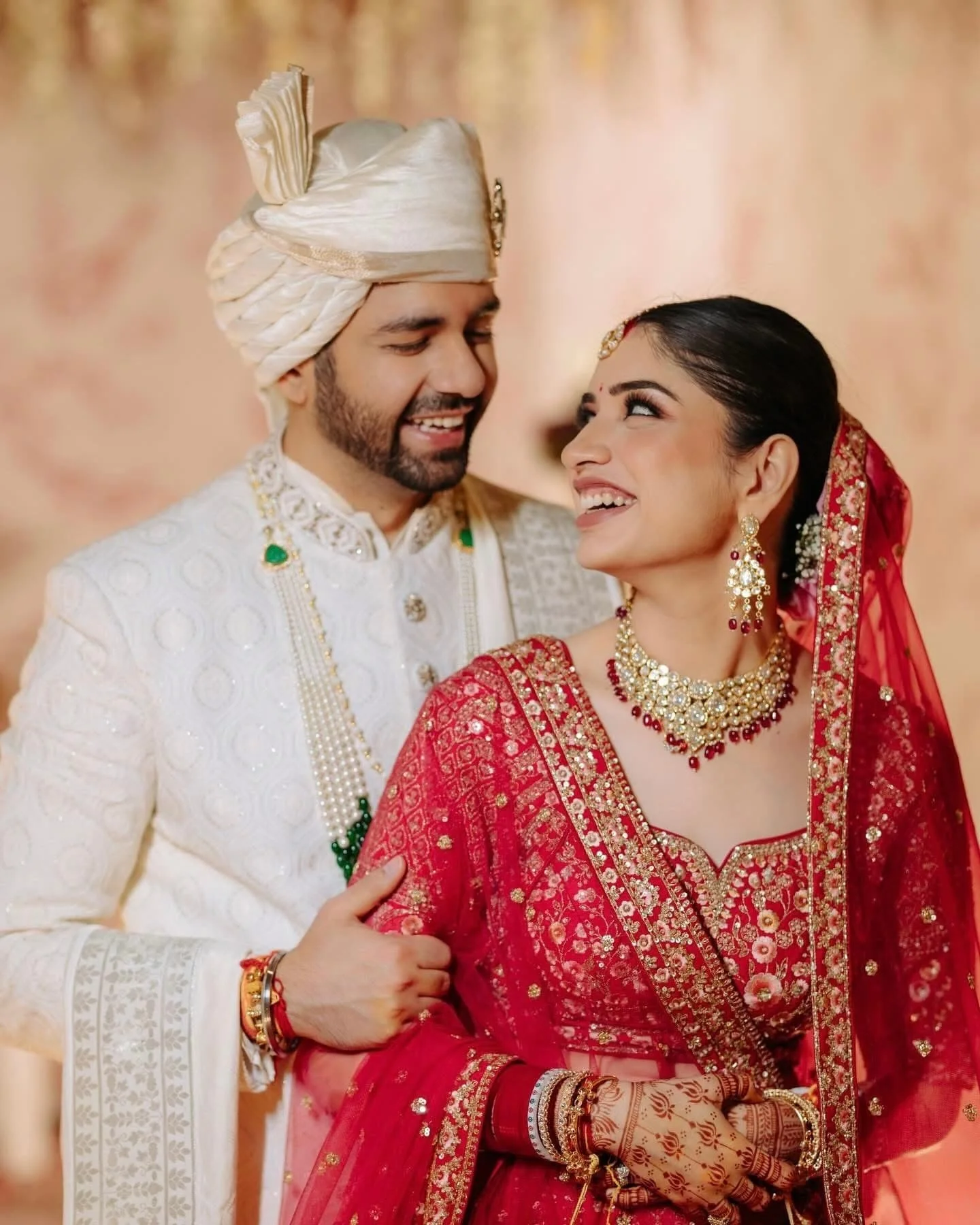 A couple in traditional Indian wedding attire smiling at each other. The man is dressed in a white embroidered sherwani and a turban, while the woman is wearing a red and gold bridal outfit with jewelry and henna on her hands.