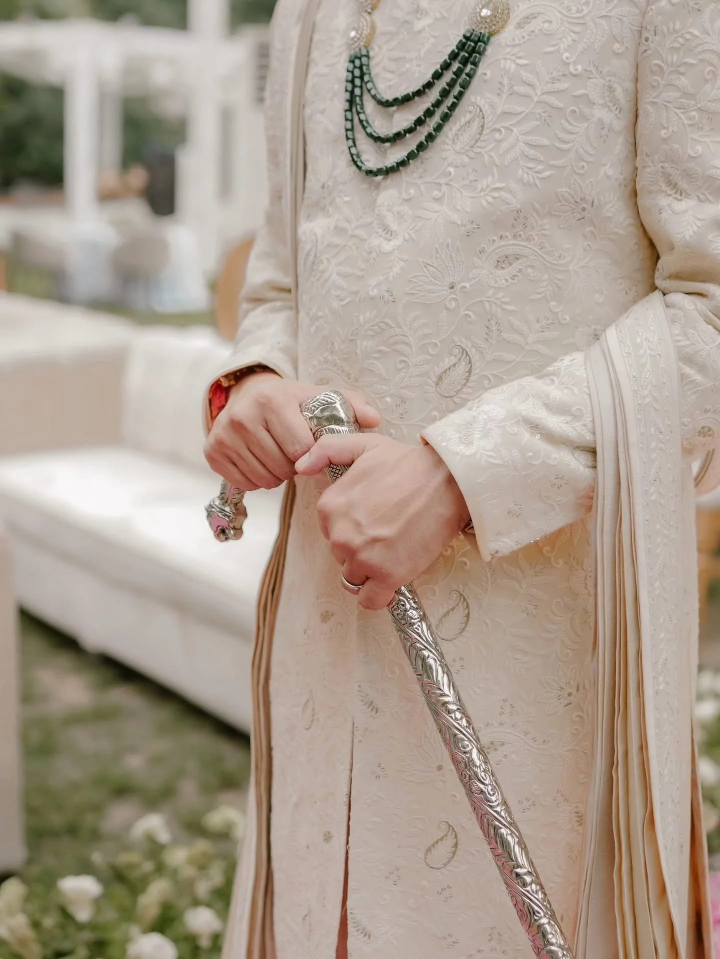 Close-up of a person in ornate, cream-colored traditional attire with intricate embroidery, holding a ceremonial staff with detailed silverwork.