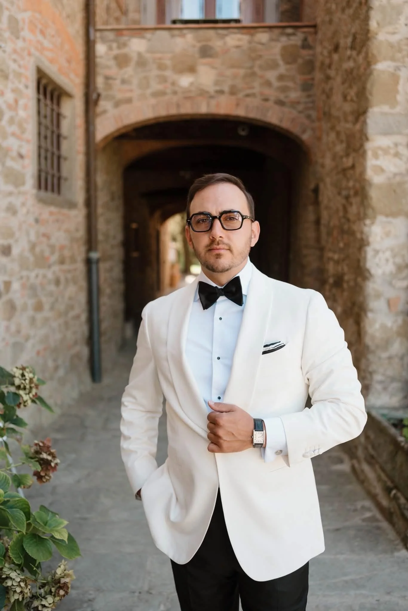 A man in a white tuxedo with a black bowtie and glasses, standing outdoors against a brick archway, with a serious expression.