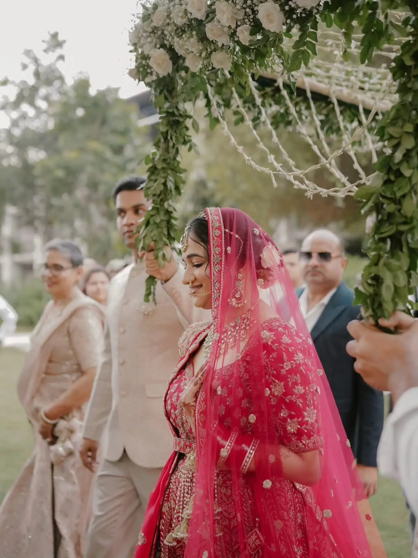 A bride dressed in a traditional pink and gold embroidered outfit with a matching pink veil, attending a wedding ceremony outdoors under a decorated floral arch, surrounded by groomsmen and other guests.