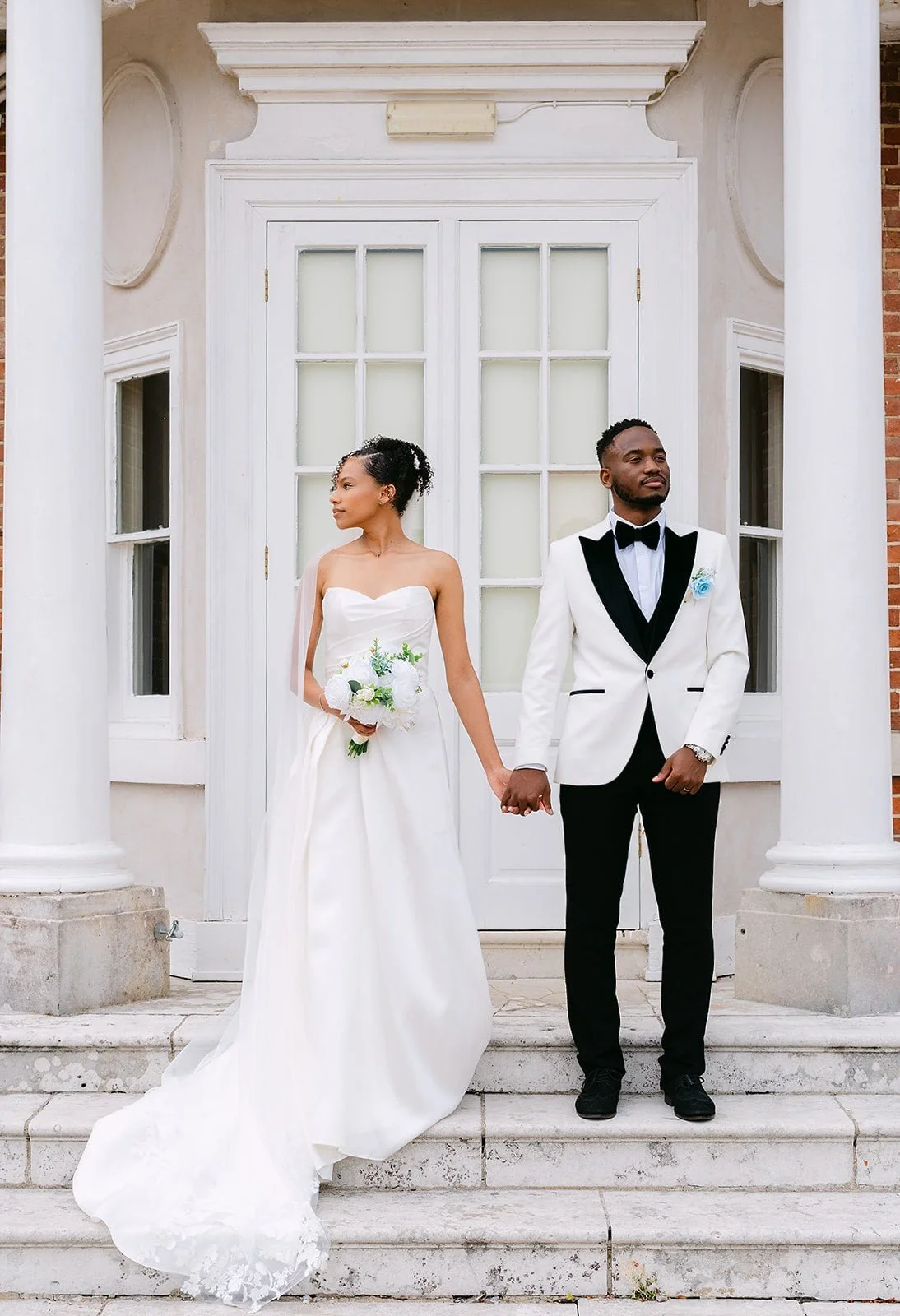 A bride and groom standing outside on the steps of a building, holding hands. The bride is wearing a white strapless wedding gown and holding a bouquet of flowers. The groom is wearing a white tuxedo jacket with black lapels, black pants, and a black