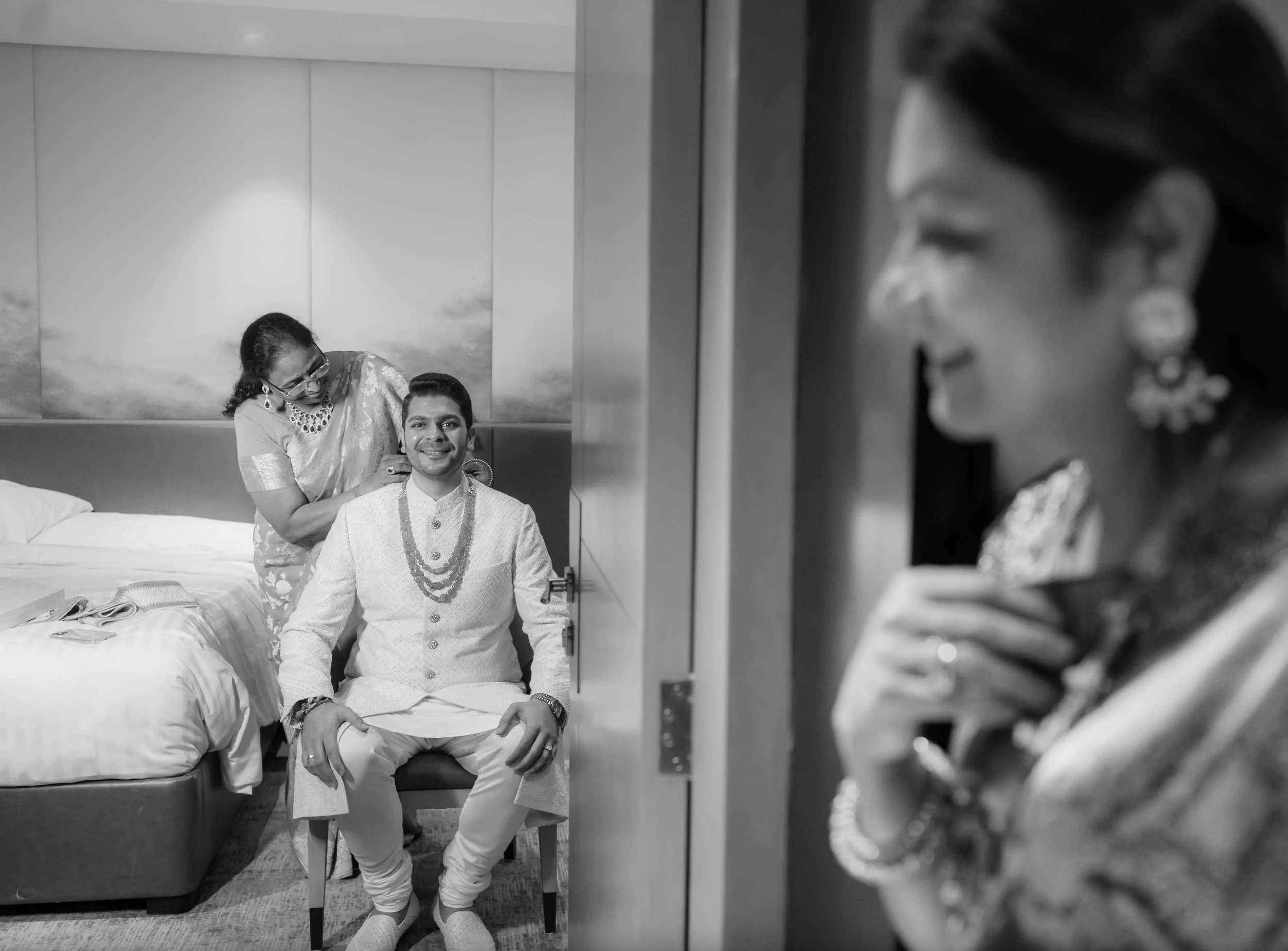 A man in traditional Indian attire sitting on a stool in a hotel room, being dressed by a woman, as seen through a mirror reflection with a woman wearing earrings and glasses in the background.