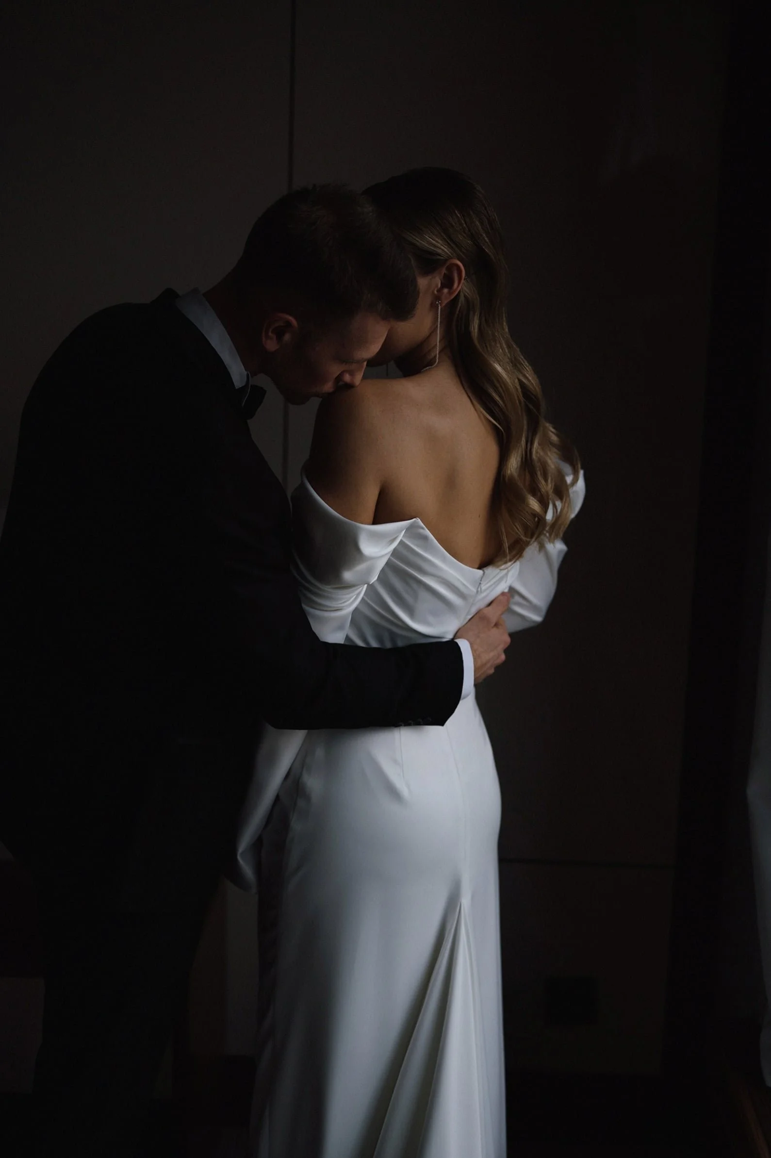 A man in a tuxedo and a woman in a white off-shoulder dress leaning in close, with the man kissing the woman's shoulder, in a dark room.
