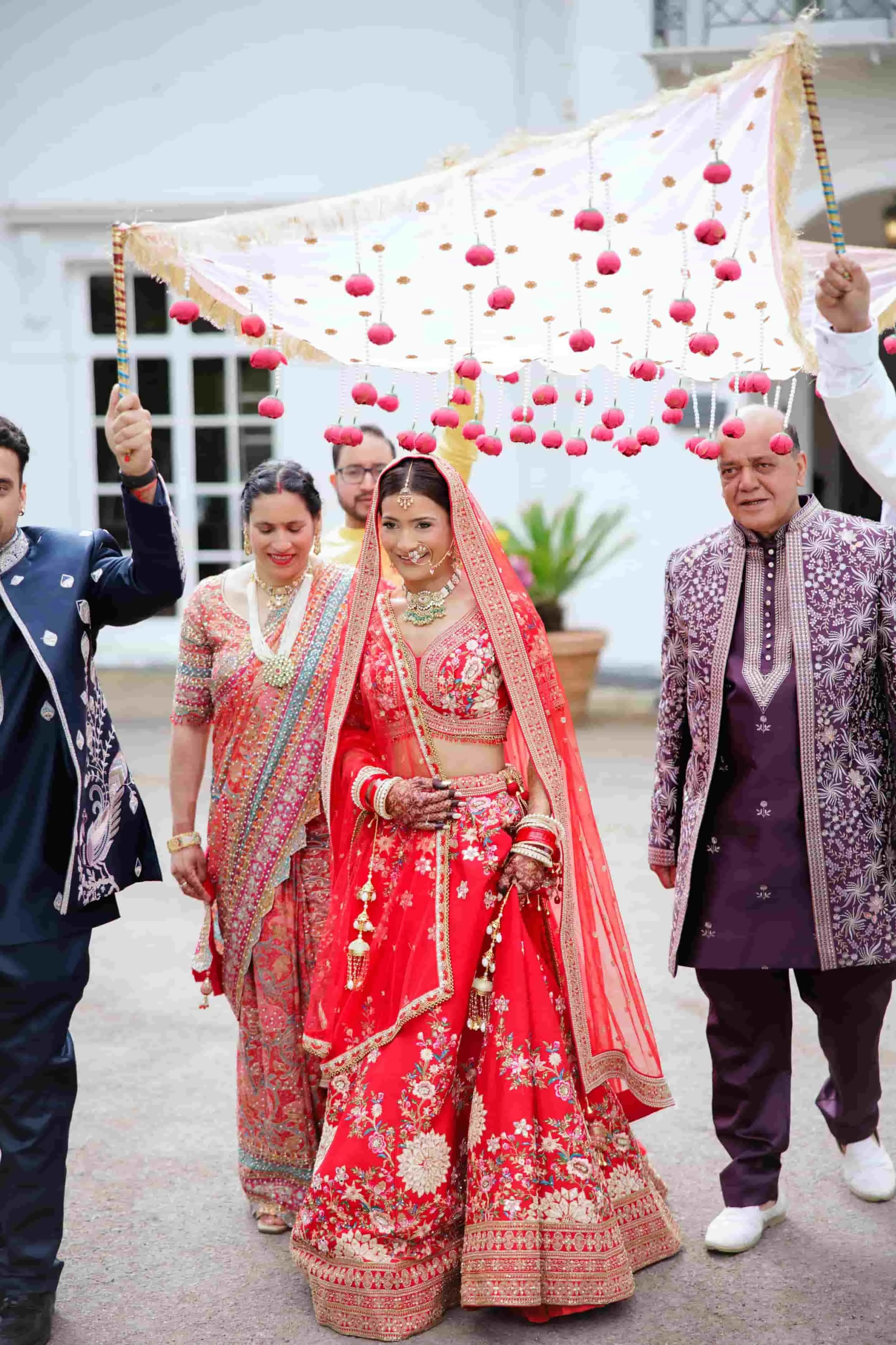 A bride in a red and gold traditional Indian wedding dress with jewelry, surrounded by her family. They are walking under a canopy decorated with pink hanging ornaments during a wedding celebration.