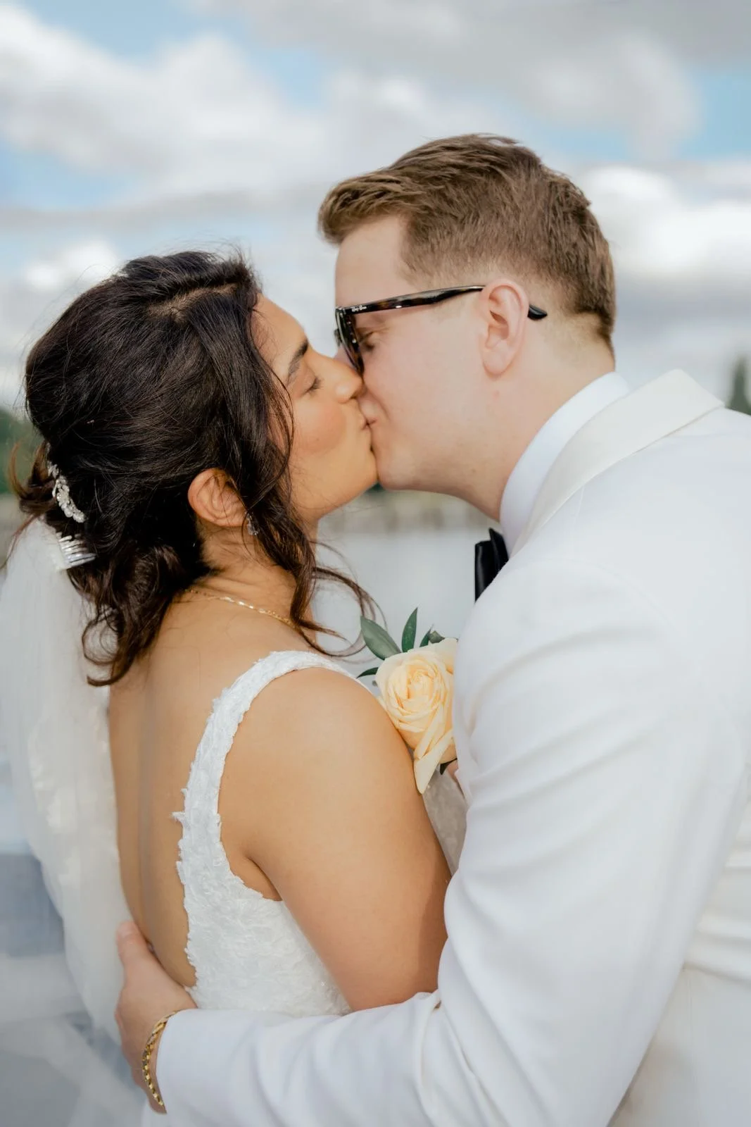 A bride and groom sharing a kiss outdoors by the water during their wedding, with the groom in a white tuxedo and the bride in a white gown holding a light-colored rose bouquet.