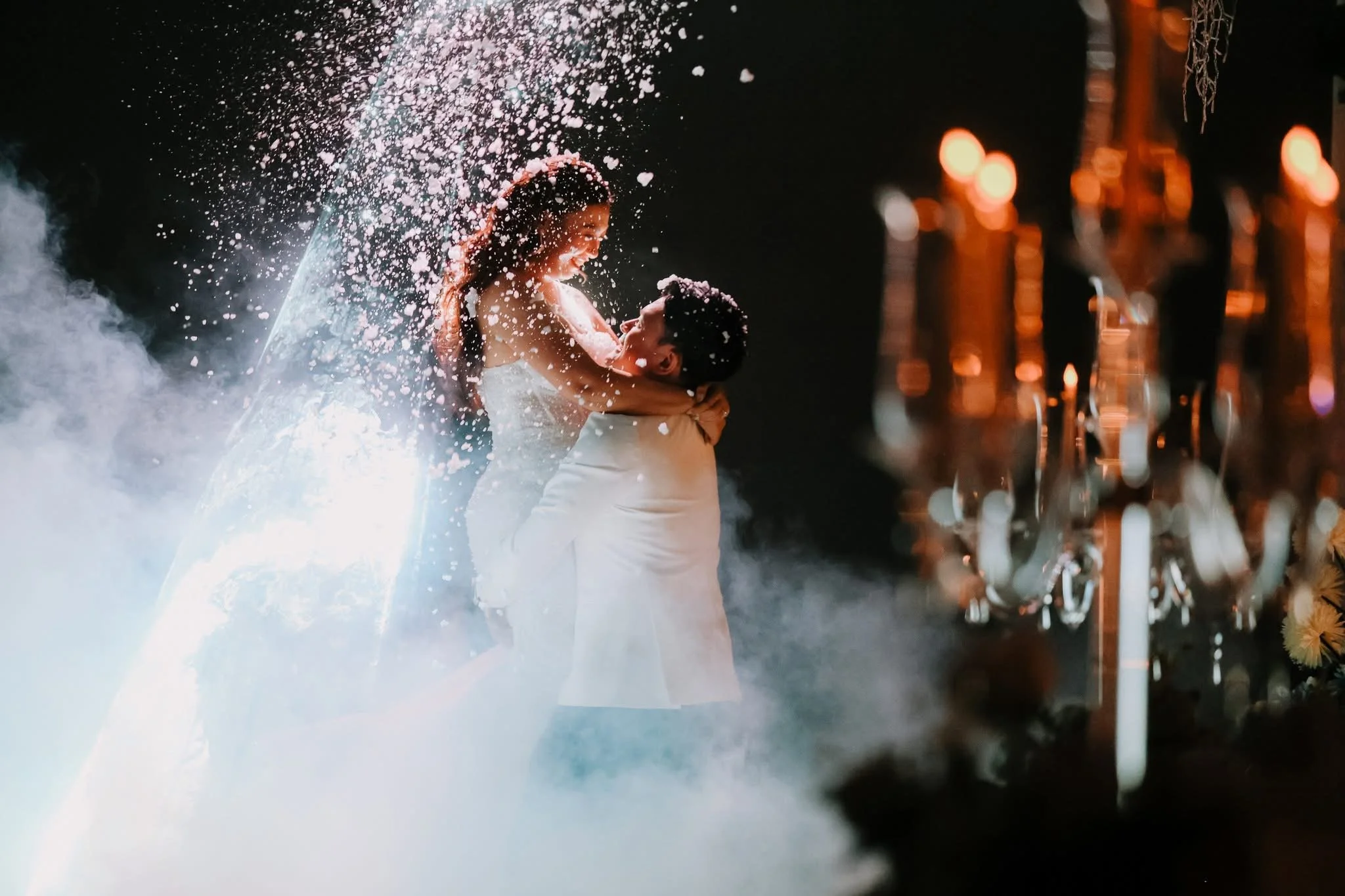 A couple embracing and smiling at each other at night with water splashing around them, illuminated by bright light, while a row of street lamps and a bridge are visible in the background.