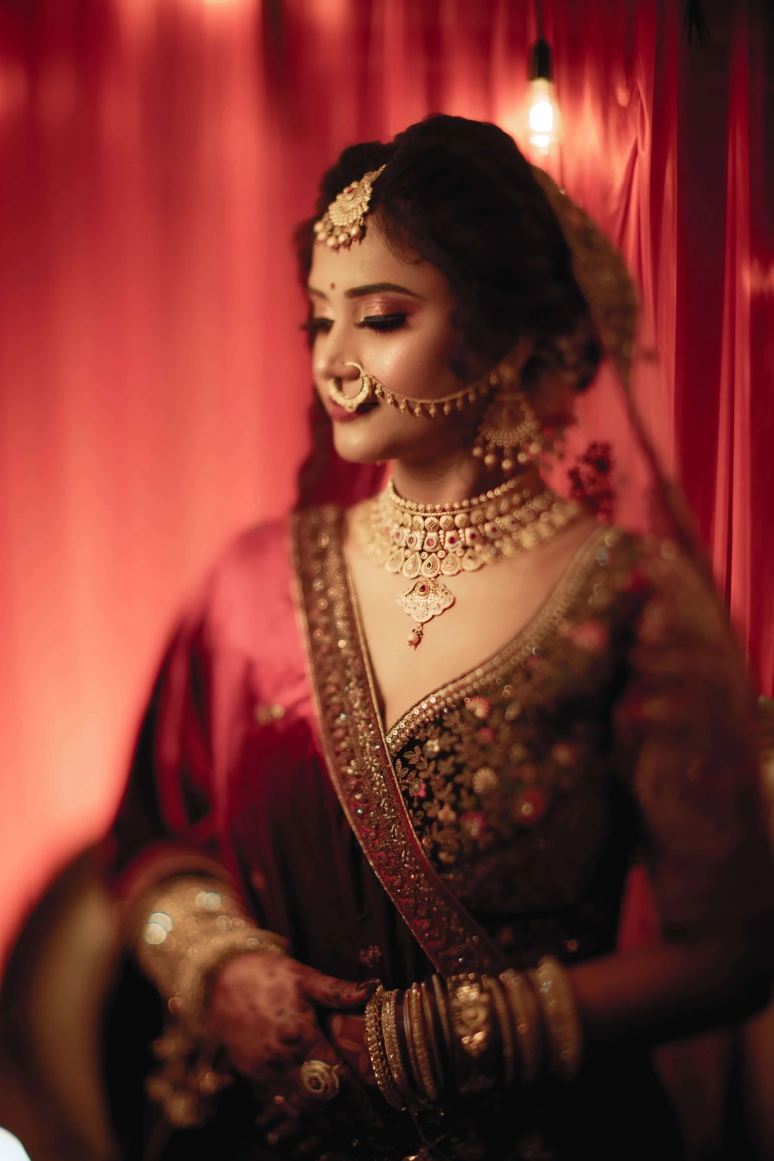 A woman dressed in traditional Indian bridal attire with gold jewelry and Bangles, standing against a red backdrop with hanging lights.