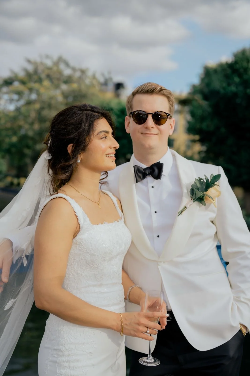 A newlywed couple dressed in wedding attire, standing outdoors, smiling, with the woman wearing a white wedding gown and veil, and the man in a white tuxedo with a black bow tie and sunglasses.